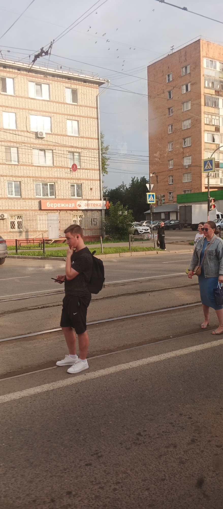 This image depicts a street scene in Izhevsk, Russia, with two residential buildings visible. The building on the left is a brick apartment complex with multiple windows and an air conditioning unit. Below it, an orange sign reads "Бережная аптека" (Beregnaya Apteka), indicating a pharmacy. The building on the right is a taller, red brick apartment building with many windows.
In the foreground, a young man wearing a black t-shirt and shorts stands on tram tracks, holding a smartphone and looking downwards. He has a black backpack on. To his right, a woman in a blue dress and denim jacket walks away from the camera, carrying a purse and a shopping bag. Another woman is partially visible behind her.
The street has tram tracks, a sidewalk, and a road with parked cars and a truck in the background. Several birds are in flight in the sky above the buildings. Pedestrian crossing signs are also visible on poles. The overall environment suggests a daytime scene in an urban setting.