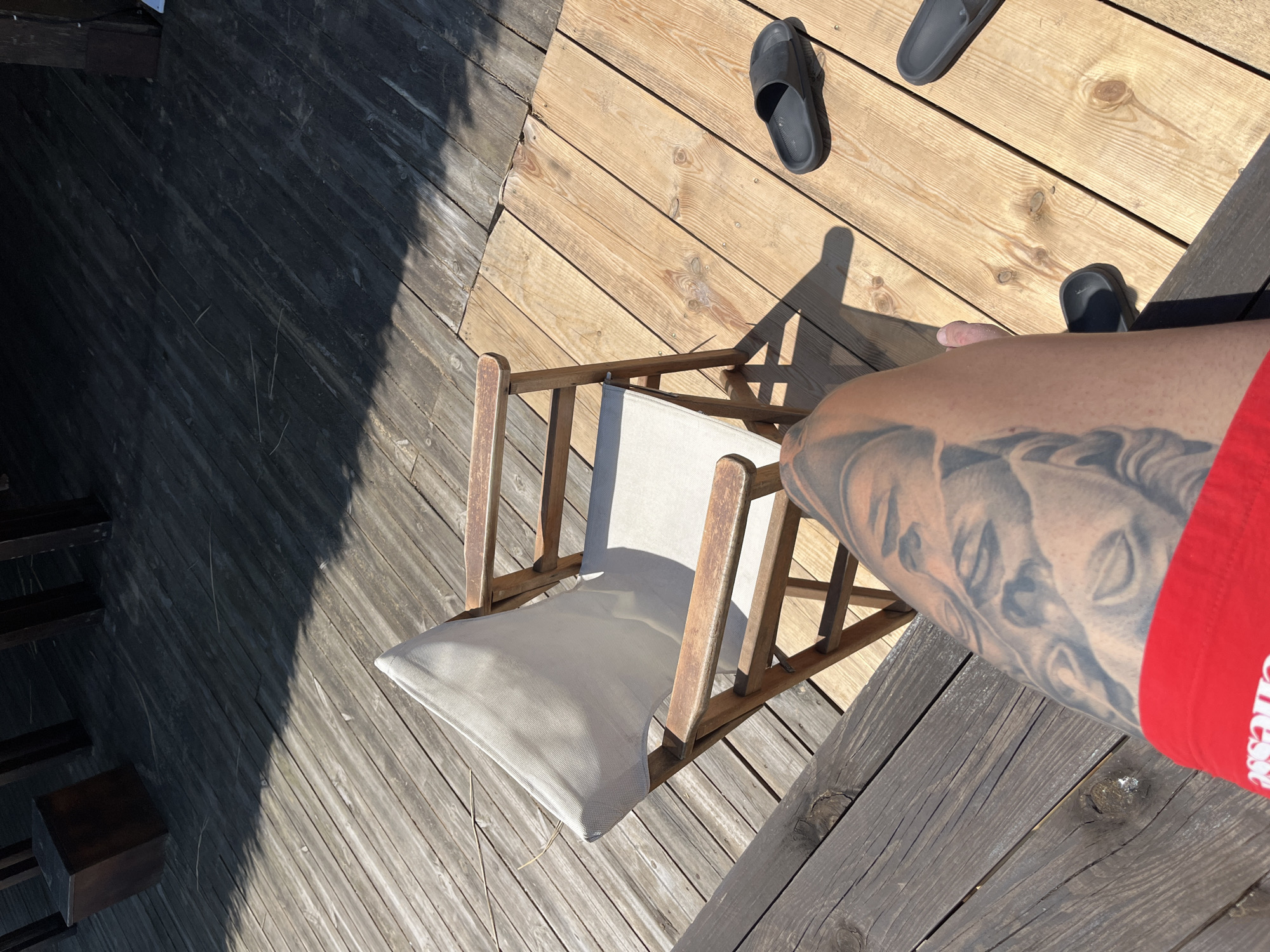 The image captures an outdoor scene on a sunlit wooden deck in Sozopol, Bulgaria. In the foreground, the upper right leg of an individual is visible, clad in red underwear or shorts that partially display the 'ellesse' logo. The leg is extensively tattooed with detailed grayscale portraits, depicting multiple classical-style human faces. A bare foot is partially visible to the right of the leg.
A wooden folding chair with a light canvas seat and back is centrally positioned, casting a sharp shadow to its right. To the right of the chair, on the light-colored wooden planks of the deck, two black rubber slide sandals are visible. The wooden deck itself shows variation in color and weathering, with distinct grain patterns and knots.
The background is dominated by deep shadow, under which dark wooden structures, possibly furniture legs or support beams, and a dark rectangular wooden box are discernible. The strong, direct sunlight creates high contrast between the illuminated areas and the shadowed portions of the scene.