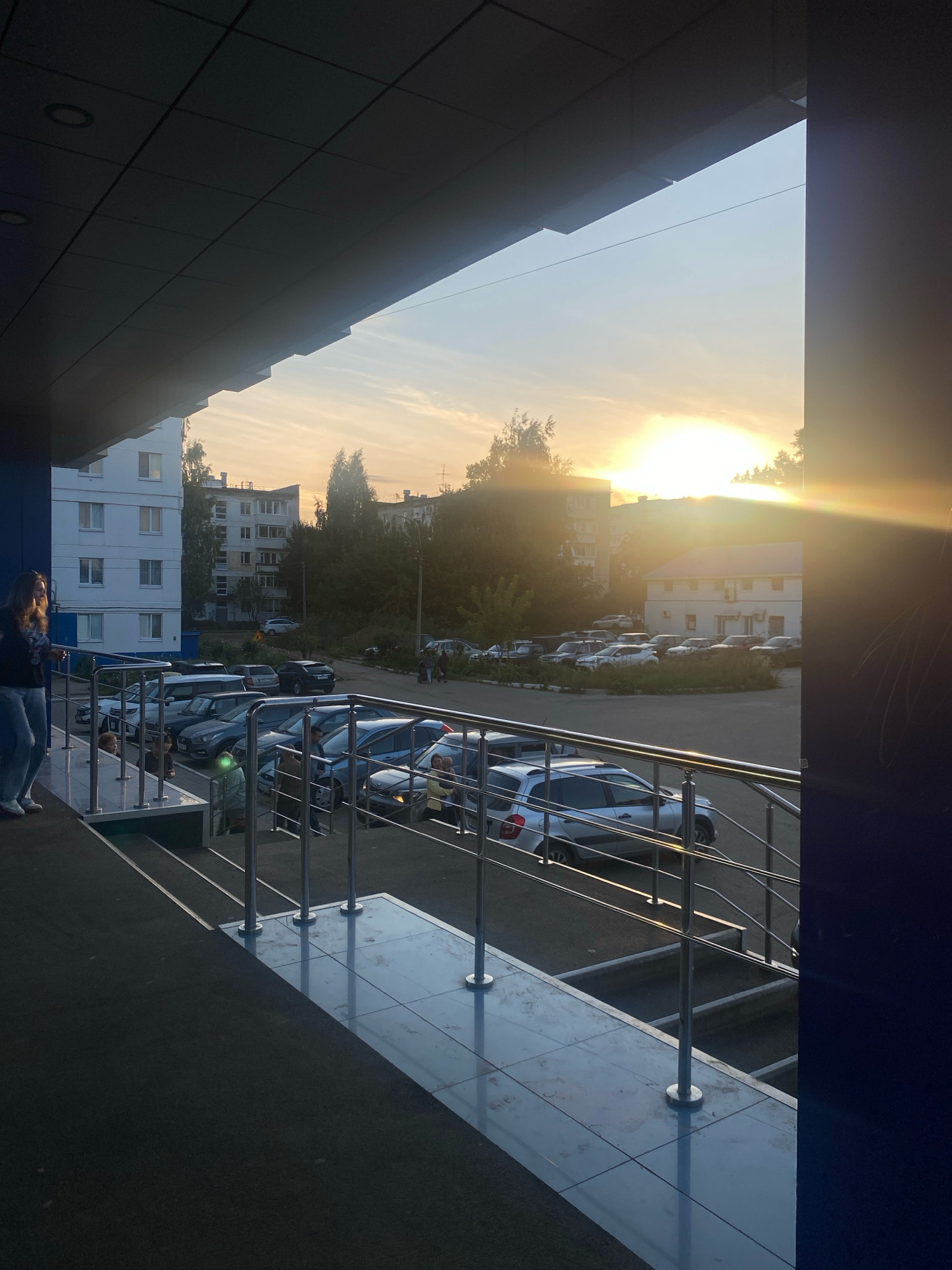The image depicts a sunset over a residential area in Chaikovsky, Russia, viewed from a covered walkway or balcony with a dark, sloped ceiling. In the foreground, a woman in blue jeans and a dark top stands near a railing, facing towards the right side of the frame. Below her, a set of stairs with a polished tiled landing and metal railings leads down. To the left, a white multi-story apartment building is visible. Across a parking lot filled with various cars, including a silver sedan and a dark SUV, are more apartment buildings and trees. The sun is low in the sky, casting a warm, golden light and creating lens flare. The overall atmosphere is that of an urban environment during early evening.