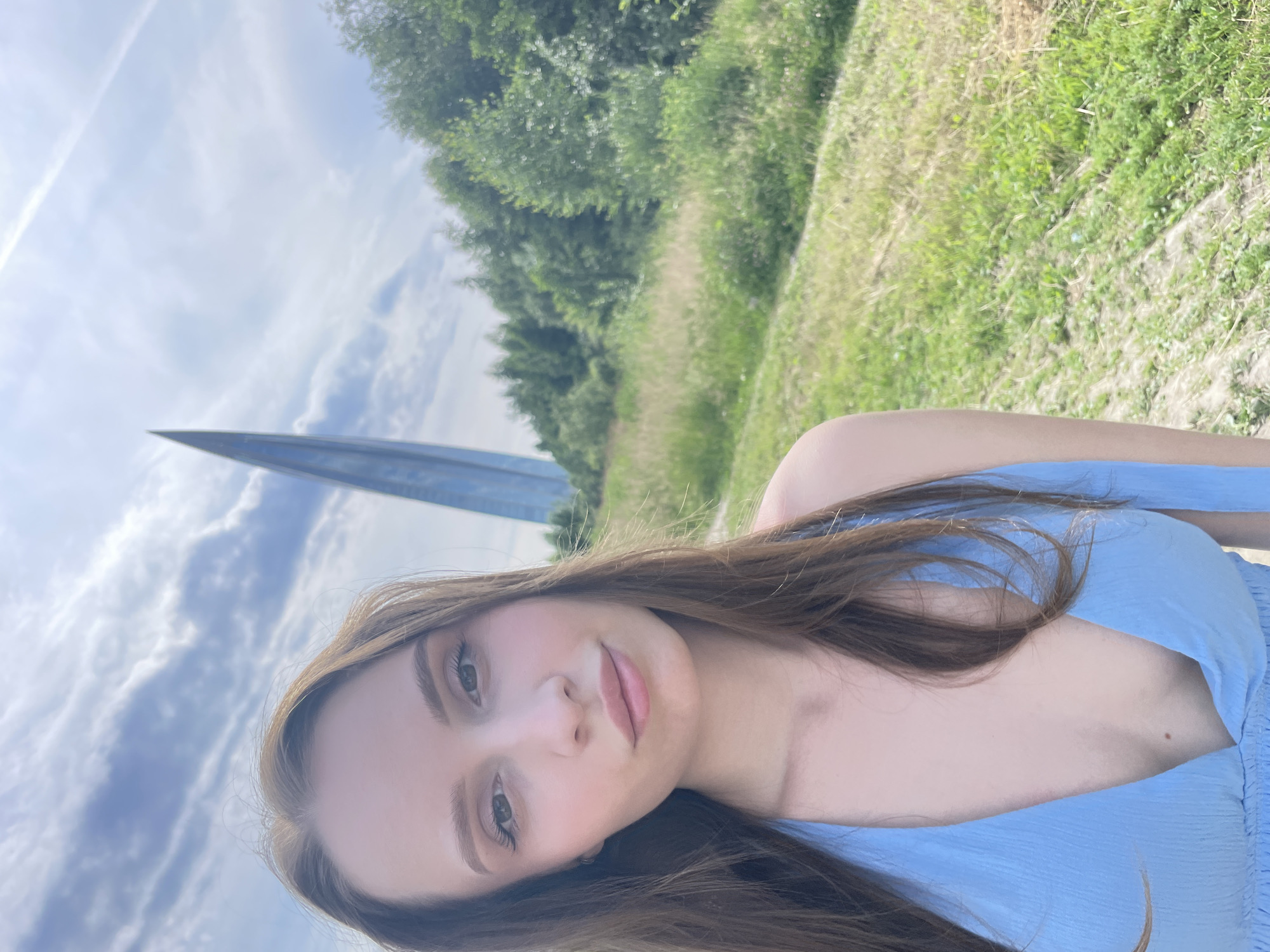 A young woman with long brown hair and a blue halter dress takes a selfie outdoors. She is positioned in the foreground, with a tall, slender, pointed building visible in the background to her left. The building is a striking, modern architectural design, reflecting the sky. The environment around her consists of a grassy field with trees in the distance under a cloudy sky. The scene is likely the area near the Lakhta Center in Saint Petersburg, Russia, an iconic skyscraper with a distinctive design.