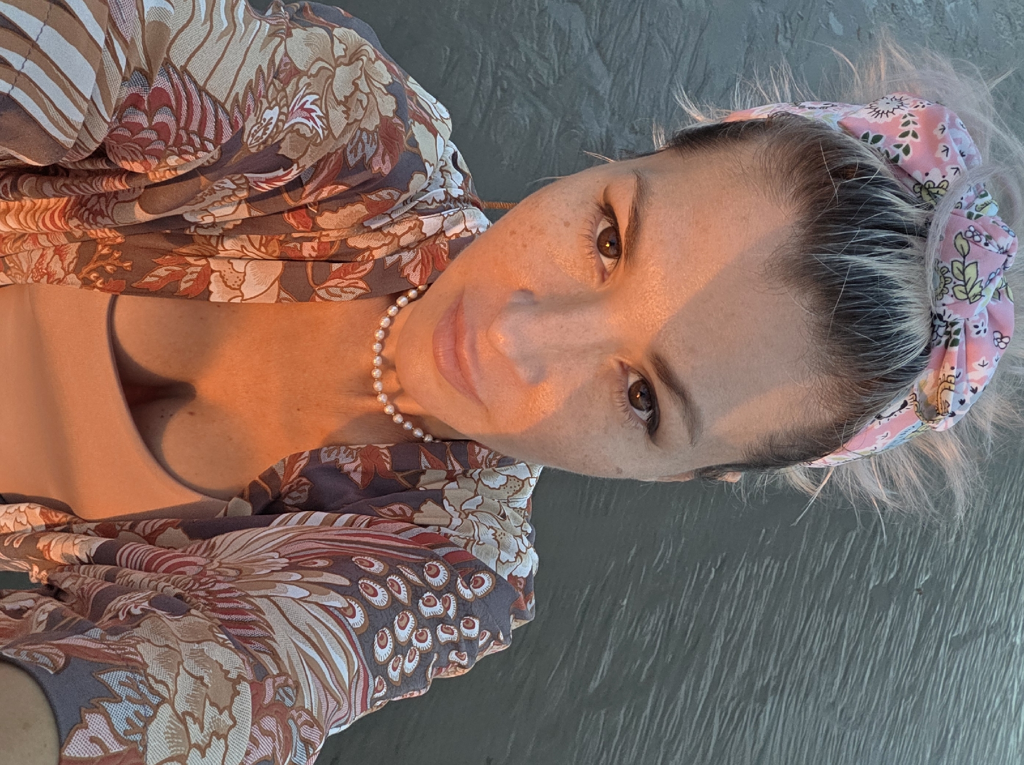 A woman with blonde hair pulled up and a pink floral headband is shown from the chest up, looking directly at the camera. She is wearing a light-colored top with a floral kimono. A pearl necklace is visible around her neck. The background appears to be a body of water, possibly the ocean, with ripples on the surface. The image is lit by warm, golden light, suggesting it was taken at sunrise or sunset. The location is identified as North Kuta, Indonesia.