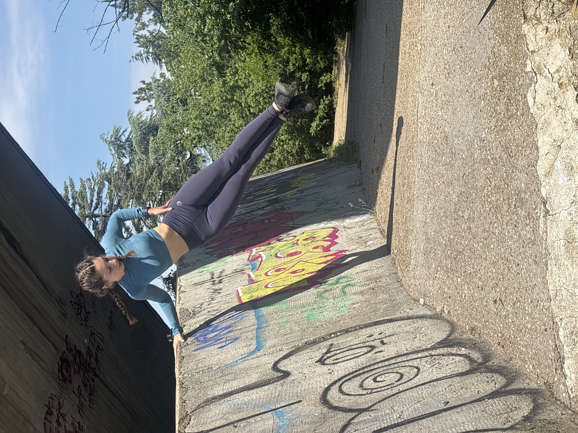The image depicts a young woman performing a horizontally extended pose against a graffiti-covered concrete retaining wall, likely situated beneath an overpass or bridge in Varna, Bulgaria. Specifically, this location appears to be an underpass or area adjacent to the Asparuhov Bridge, known for its concrete structures and street art.
The woman, with braided brown hair, wears a teal long-sleeve crop top, dark navy leggings, and black athletic shoes. She is positioned with her left hand pressed against the concrete wall for support, her torso angled downwards, and her legs extended horizontally, parallel to the ground, with her right hand resting near her hip. She looks down and to her left.
The concrete wall features various graffiti, including a prominent yellow and pink abstract design, black outlines, and blue and green markings. Above the woman, the grey underside of a concrete bridge or overpass is visible, also bearing faint graffiti. The ground is a light grey, pebbled concrete path with some cracks. In the background, lush green deciduous and coniferous trees fill the midground under a clear blue sky, with a dirt path winding through them. Strong sunlight casts distinct shadows on the wall and ground.