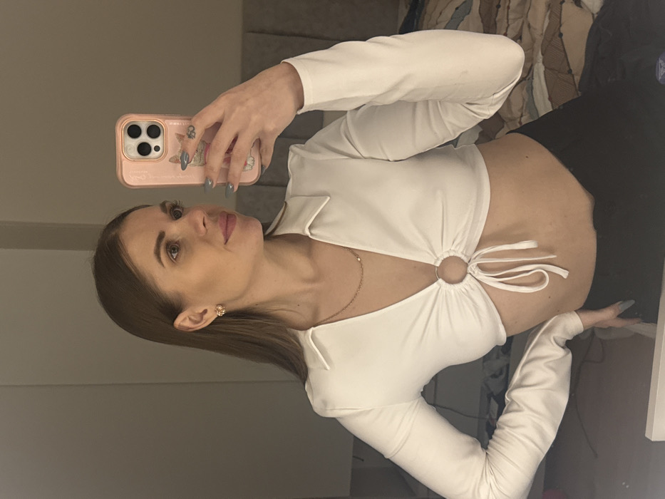 A woman is captured in a selfie, her phone prominent with a pink case. She's wearing a white, long-sleeved crop top with a distinctive ring detail at the chest. Black pants are visible. Her hair is brown and styled. She has earrings. The background includes a mirror and a glimpse of a patterned surface, possibly a bed or couch. The lighting is soft, highlighting her features and attire.