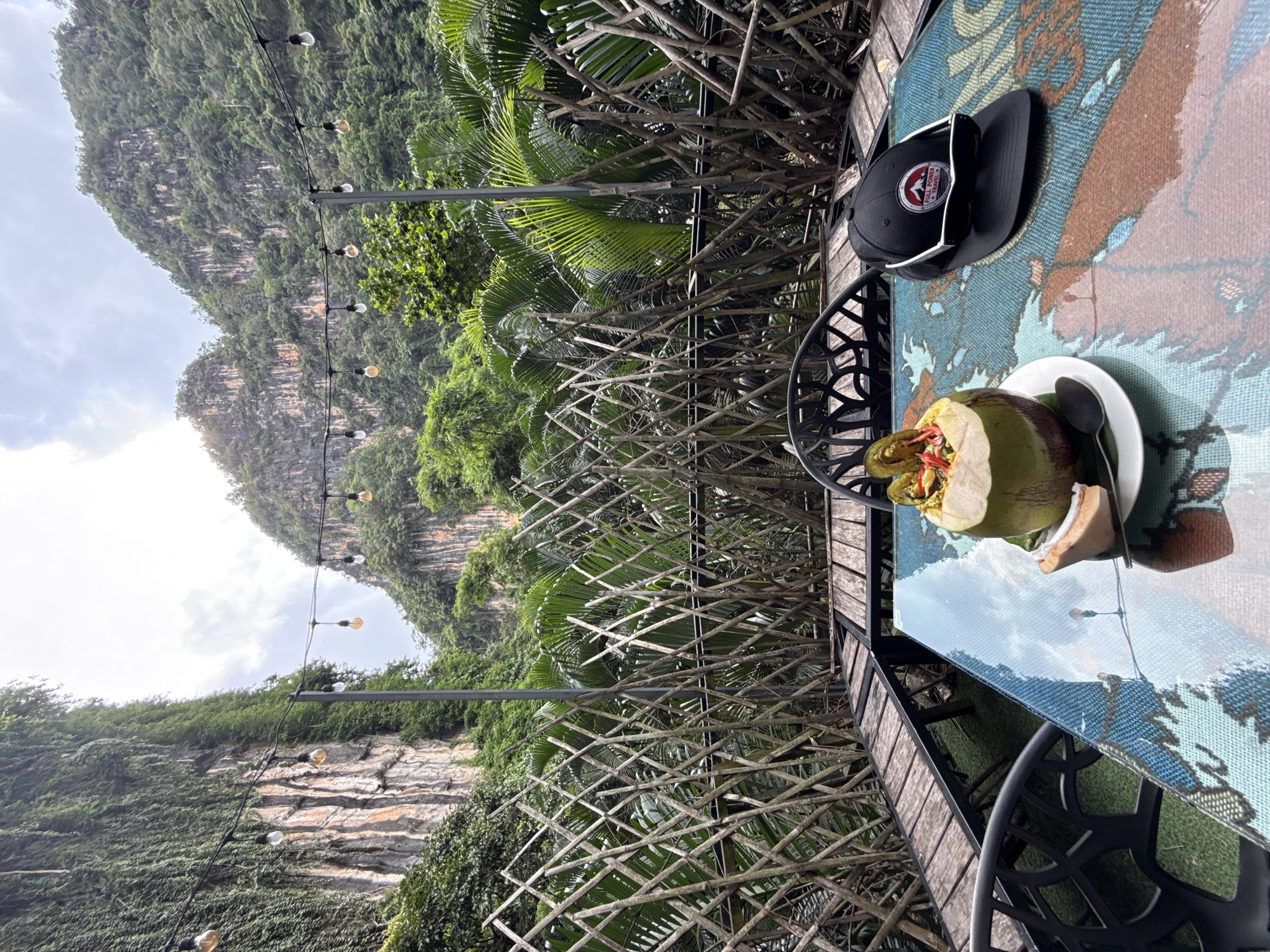 In Chong Pli, Thailand, a table at an outdoor restaurant displays a halved coconut filled with a colorful fruit salad, a slice of coconut on a plate with a spoon, and a black baseball cap with sunglasses resting on top. The table has a patterned glass top reflecting the surroundings. Behind the table, a woven bamboo fence separates the seating area from lush tropical foliage and towering limestone cliffs draped with green vegetation. String lights are suspended across the scene, and the sky is overcast.