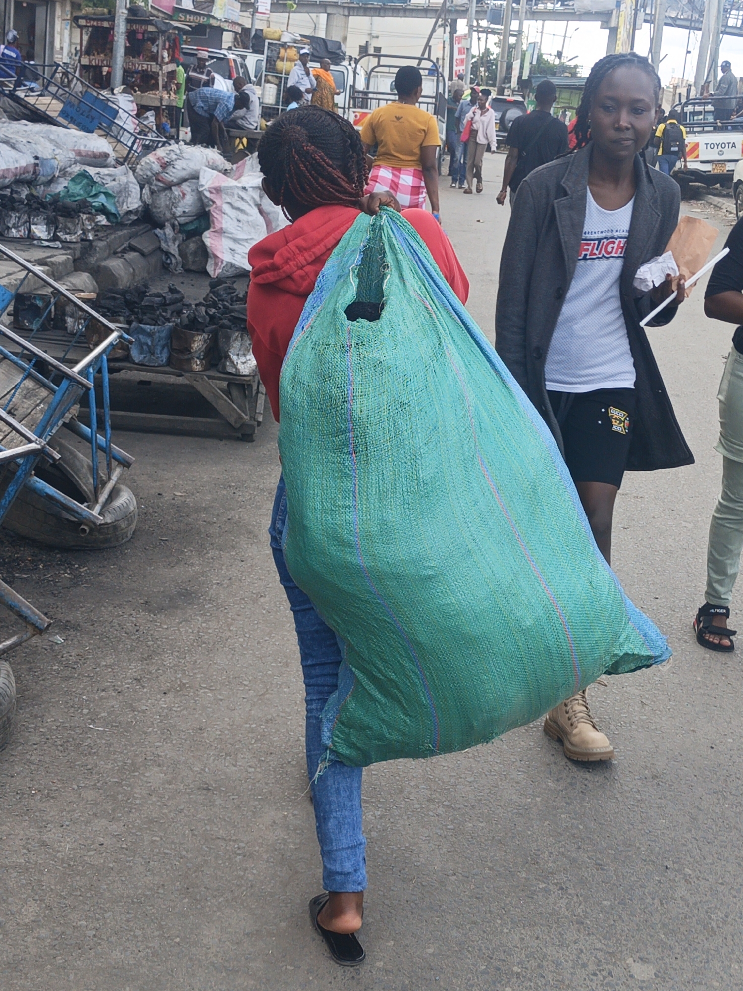 A medium shot shows two young African women in the city of Nairobi, Kenya. One woman faces away and carries a large, full, blue-green sack over her right shoulder. She is wearing a red hooded sweatshirt, blue jeans, and black sandals. The second woman faces the camera. She wears a gray blazer over a white tee shirt, black shorts, and beige work boots. She carries papers and a white disposable stick in her left hand. Behind them, multiple people walk along the roadside. Cars are parked in the background, including a white Toyota truck. To the left of the carrier, overturned blue metal carts and sacks of goods sit near what appears to be charcoal. Overhead powerlines stretch across the image.