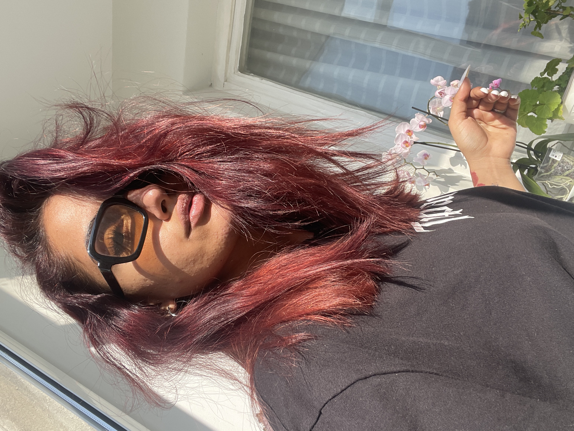 A person with dark red hair and wearing black-framed glasses is positioned near a window in an indoor environment. The person is wearing a black shirt. The person's left hand is holding a pink orchid flower. A potted plant is visible on the windowsill next to the orchid. Sunlight is streaming through the window, creating shadows on the person's face and hair. The location is Unknown, Unknown.