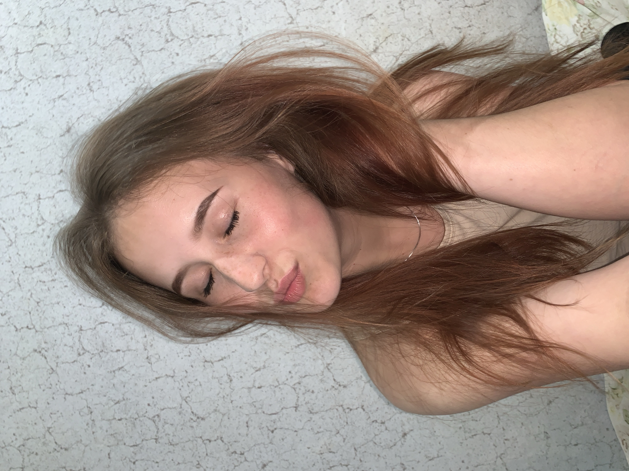 A close-up, slightly overhead shot depicts a young person, likely female, lying down with her eyes closed, appearing to be asleep or deeply relaxed. Her head is tilted slightly to the right, and her long, voluminous reddish-brown hair is spread out, framing her face and flowing onto the surface she is resting on. She has a fair complexion with a scattering of light freckles visible across her nose and cheeks. Her eyebrows are neatly shaped, and she has applied winged eyeliner. Her lips are naturally full and slightly parted. She wears a light-colored, possibly beige, top, and a slender silver hoop earring is visible in her right ear. The background consists of a light grey or off-white textured surface, featuring a subtle, crackle-like pattern, suggesting an indoor setting such as a bed or floor. A small portion of a floral patterned fabric is visible in the upper right corner, possibly a pillow or blanket. The overall impression is one of peaceful rest. No visible text is present. The location context for this image is Фоки, Russia.