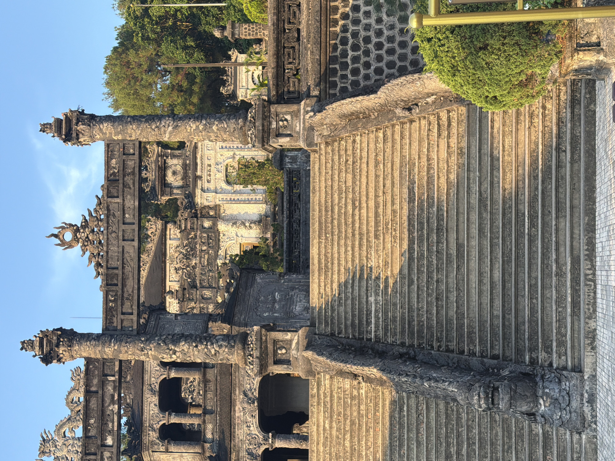 The image depicts a stone architectural structure in Thủy Xuân Ward, Vietnam. The structure features a prominent stairway with vertically ribbed steps, flanked by ornate stone pillars and walls. The pillars are intricately carved with dragon motifs and geometric patterns. Behind the structure, there's a glimpse of another white-and-blue decorated building, suggesting the structure is part of a larger complex. Vegetation and trees are visible in the background under a clear blue sky. The lighting suggests it's either early morning or late afternoon.