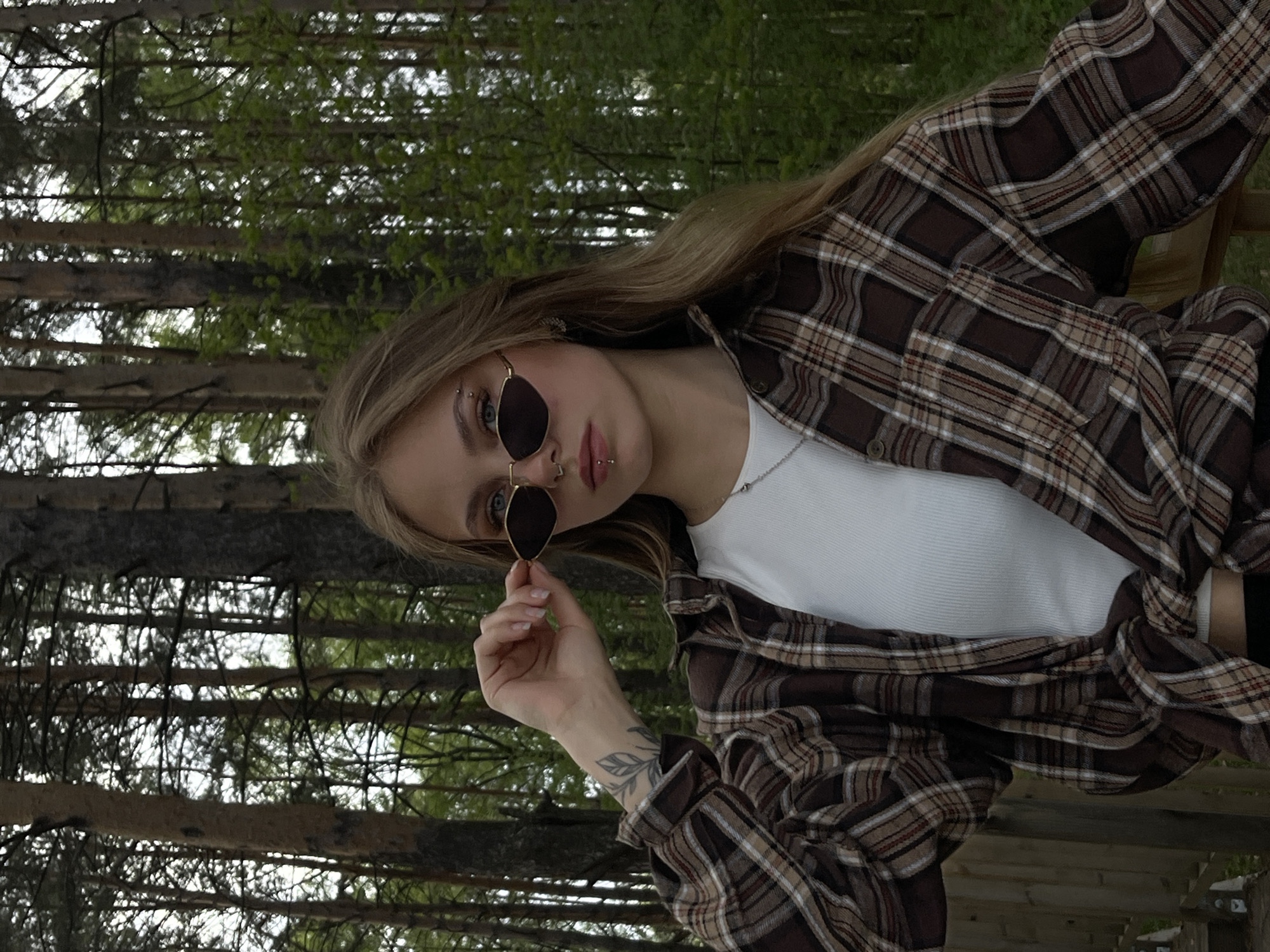 A young woman stands outdoors, appearing to be in a wooded area. She is wearing a brown plaid shirt over a white top, and is holding sunglasses. She has blonde hair and facial piercings. The background shows trees, suggesting a forest or park setting. There is no explicit indication this image was taken in Chaikovsky, Russia.