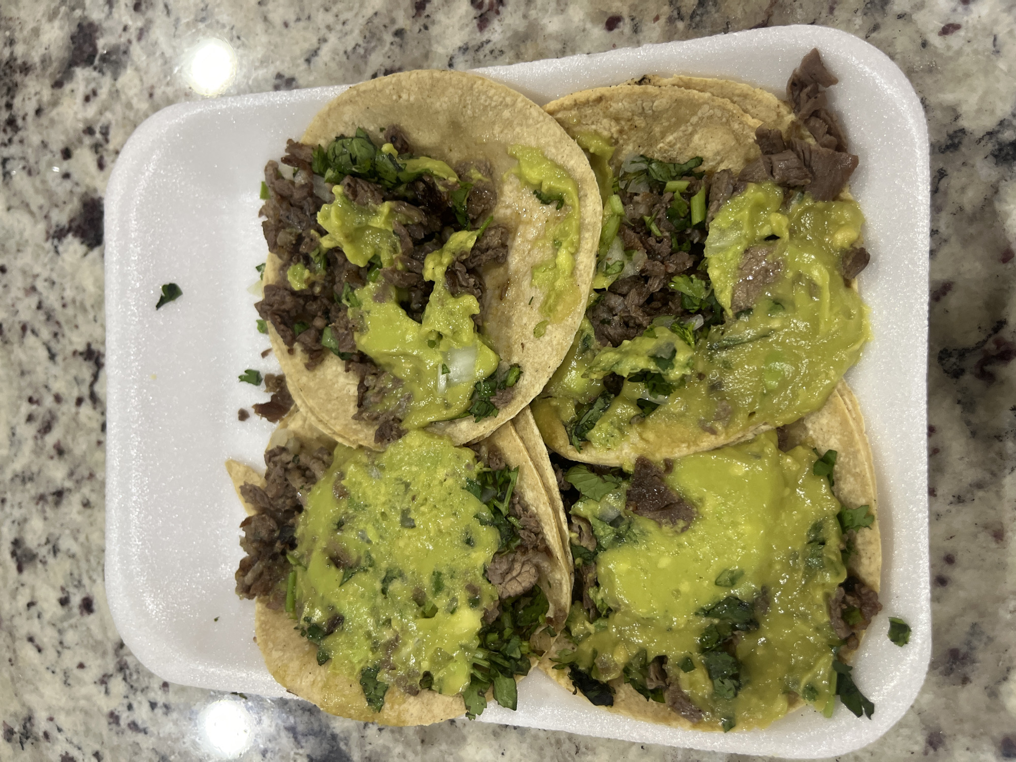 The image depicts four street tacos in a white, rectangular styrofoam container on a beige countertop with a speckled pattern. Each taco contains carne asada, cilantro, and what appears to be guacamole or avocado sauce. The tacos are arranged closely together within the container. The scene suggests a simple, informal setting, potentially a takeout meal. There is no explicit information to confirm the exact location as Barrio de Nuevo León, Mexico; this is an unsupported detail. No people are visible in the frame.