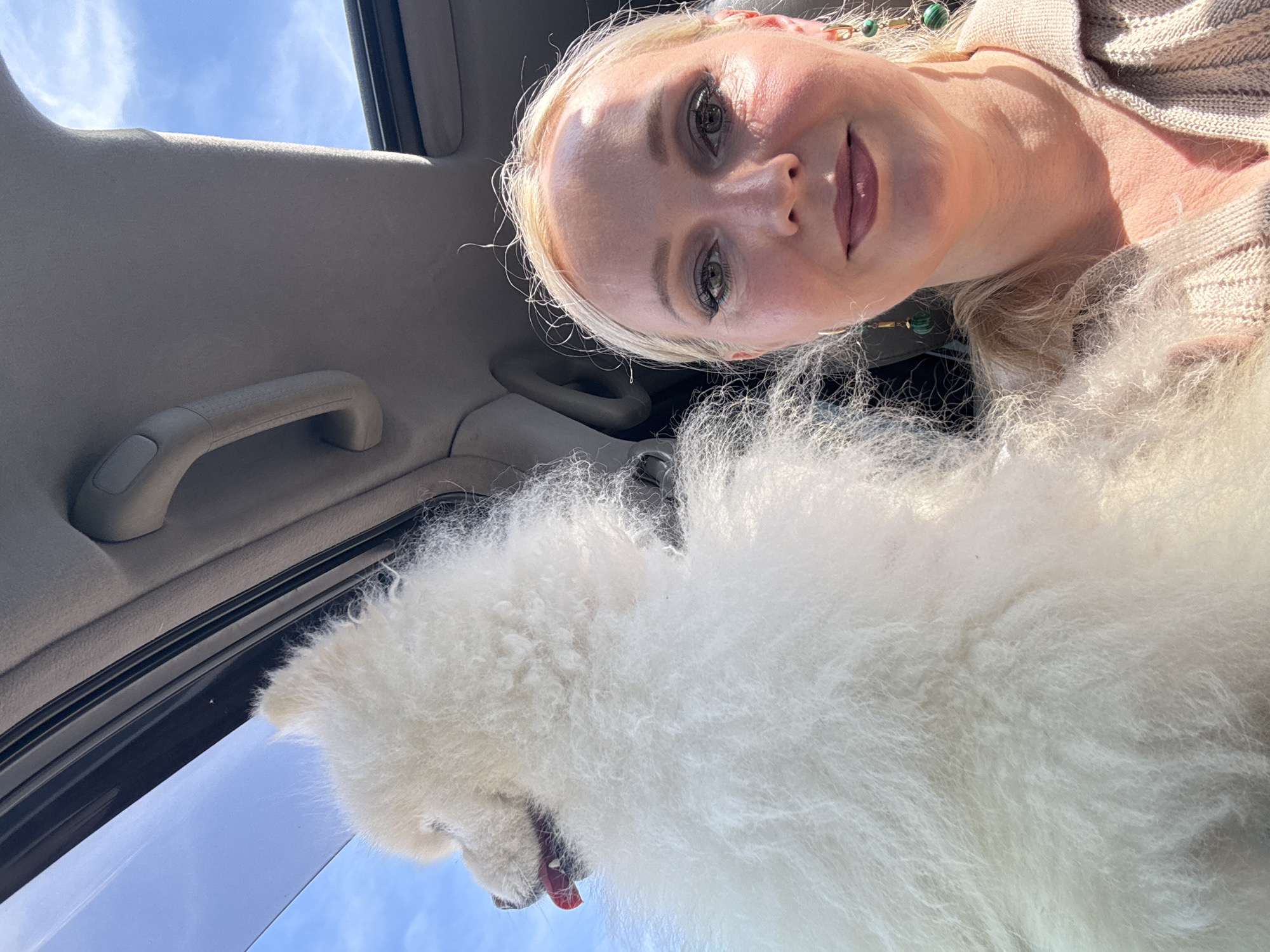 A woman with blonde hair and makeup smiles at the camera from inside a car. Her left side is partially obscured by a large, fluffy white dog that is looking out the open sunroof. The car's interior is visible, including the headliner and a grab handle. The sky is blue with scattered clouds. The location is not identifiable as Komsomolsk-on-Amur, Russia from the visual information provided.