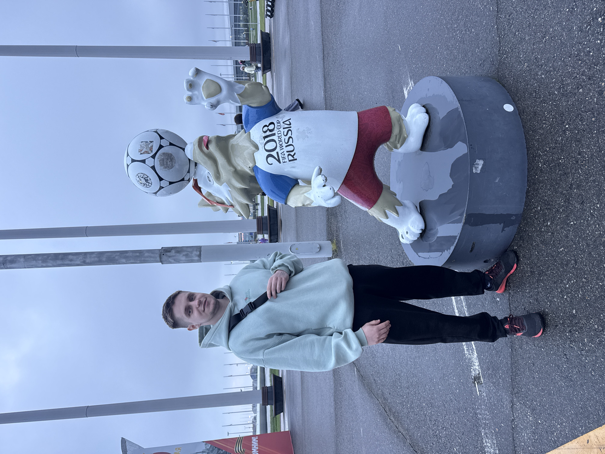 A man in a light green hoodie and black pants stands next to a mascot statue on a gray paved area in Sirius, Russia. The mascot holds a soccer ball and wears a shirt with "2018 FIFA WORLD CUP RUSSIA" printed on it. In the background, there are metal poles, benches, and other people. The weather appears overcast.