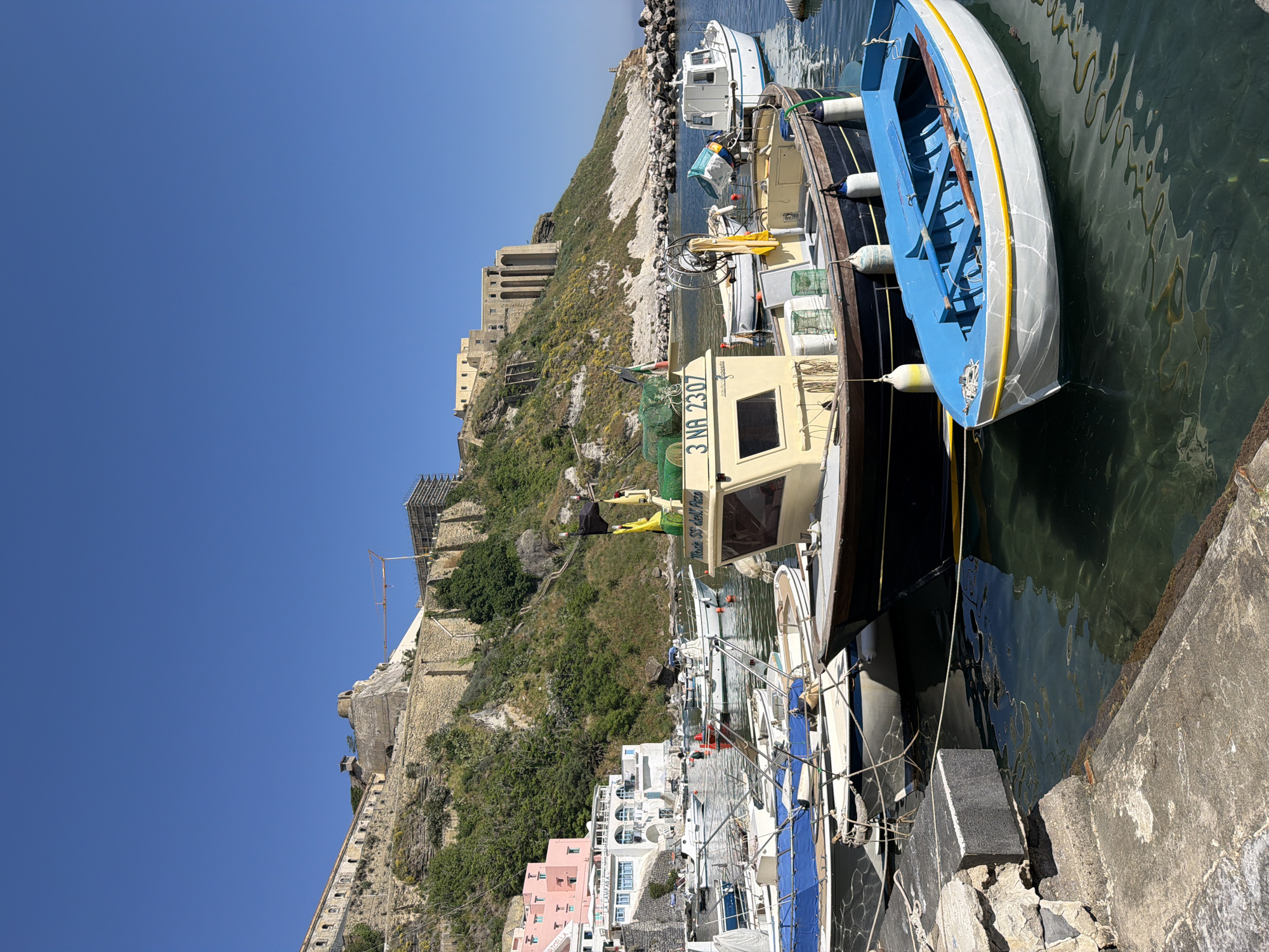The image shows a harbor scene in Procida, Italy, with several boats docked along a concrete pier. A blue and white rowboat is visible on the right side of the frame, tied to a larger boat. The larger boat is painted in tan and black with the registration number "3 NA 2307." In the background, a steep hillside rises, topped by a historical fortress with scaffolding and construction equipment visible. Buildings in shades of white and pink are clustered at the base of the hill. The sky is clear and blue. The water in the harbor reflects the light and shadows of the boats and surrounding structures.