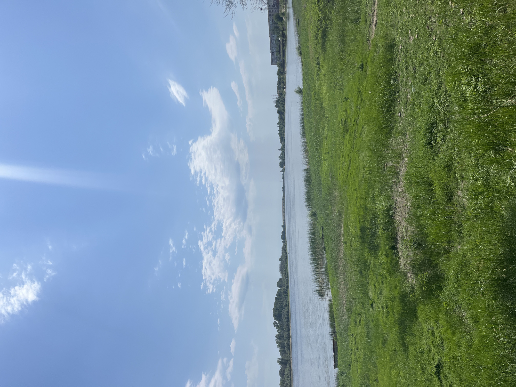 The image depicts a wide, calm river with a lush green grassy bank in the foreground. The river stretches across the frame, with a treeline visible on the opposite bank. In the distance, on the right side of the river, a multi-story building complex can be seen. The sky is bright blue with scattered white clouds, and a distinct sunbeam cuts through the upper left portion of the frame. The grassy bank is slightly uneven with some patches of dry earth visible. Two small, weathered boats are partially submerged at the water's edge on the left side of the image. The scene appears to be a tranquil, natural environment within a populated area.
