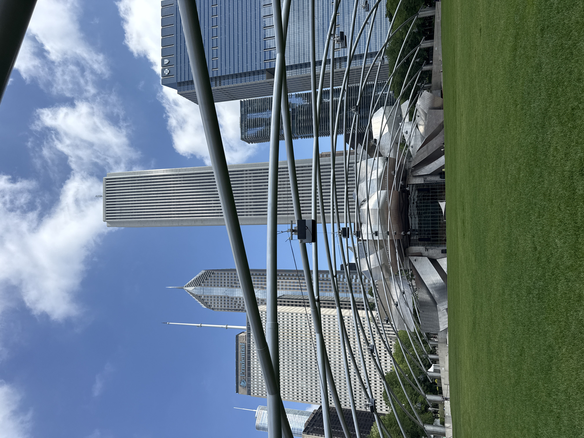 This image shows a view of Jay Pritzker Pavilion in Millennium Park, Chicago, Illinois, United States of America. The pavilion's distinctive stainless steel canopy structure dominates the foreground, with its parabolic arches curving over a large grassy lawn. In the background, several tall skyscrapers rise against a partly cloudy blue sky.
The pavilion itself features a stage area with a metallic, sculptural facade. Several speaker boxes are suspended from the canopy's framework. The lawn in front of the pavilion is lush and green, appearing well-maintained. A few people are visible in the distance on the lawn and near the pavilion, appearing to be casually walking. The overall scene depicts a public urban park space on a bright, clear day.