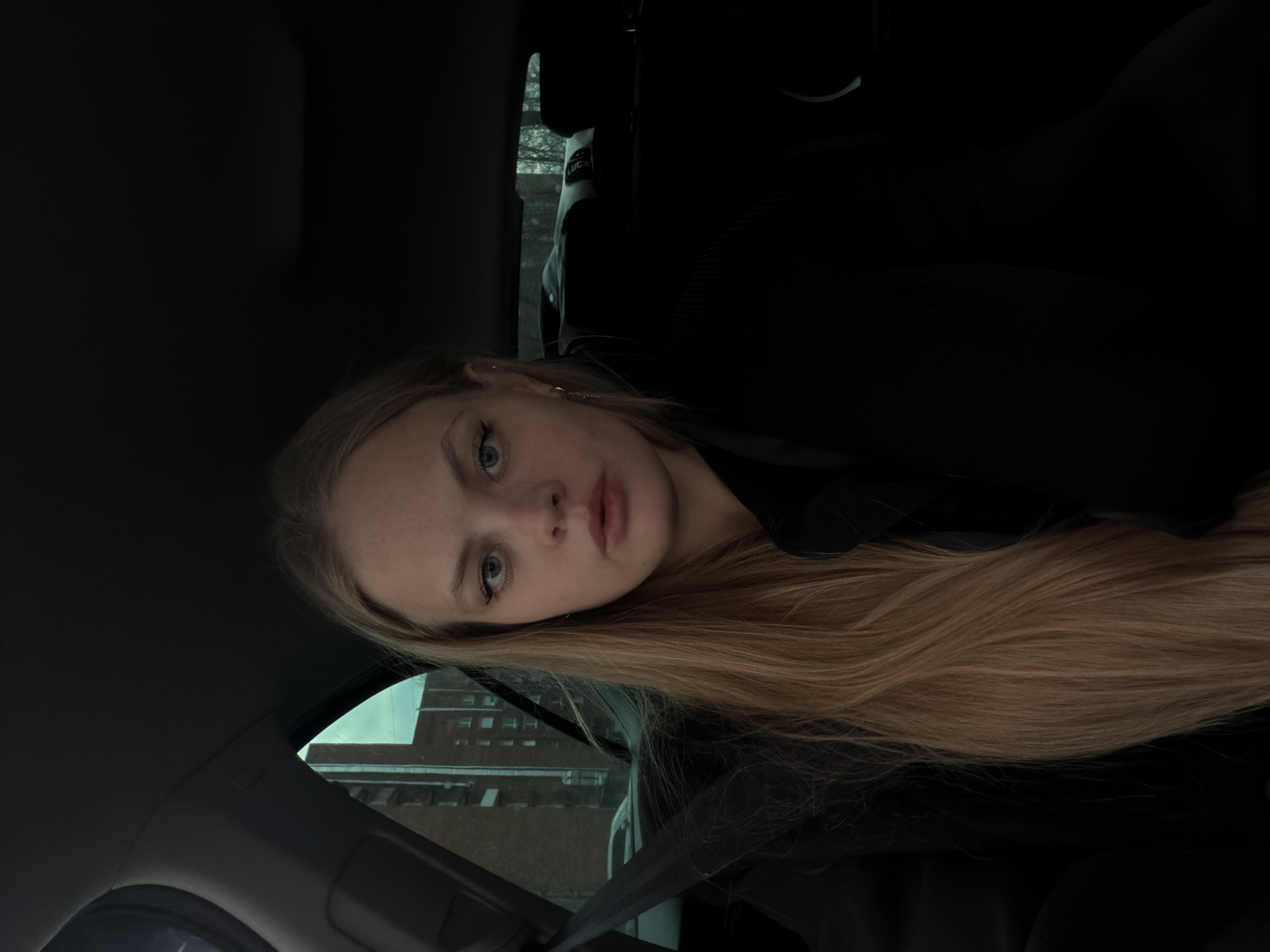The image shows a young woman with long, light blonde or light brown hair and light-colored eyes, positioned in what appears to be the passenger seat of a car. She is looking directly at the camera with a neutral expression, her lips slightly parted. A seatbelt is visible across her chest.
The scene is set inside a car, characterized by dark interior elements including the headliner, seats, and a visible headrest in the background. Through the car window on the left, a large, dark-colored building with multiple rectangular windows is visible, suggesting a brick or concrete structure. The light coming through the window indicates it is daytime, but the overall dimness and muted tones suggest an overcast sky or low light conditions. Hints of bare trees or vegetation can be seen between the building and the car.
The woman is wearing dark clothing. On an object in the background, possibly a headrest or a bag, faint letters "LUC" are partially visible. The location context provided indicates this scene is in Izhevsk, Russia.