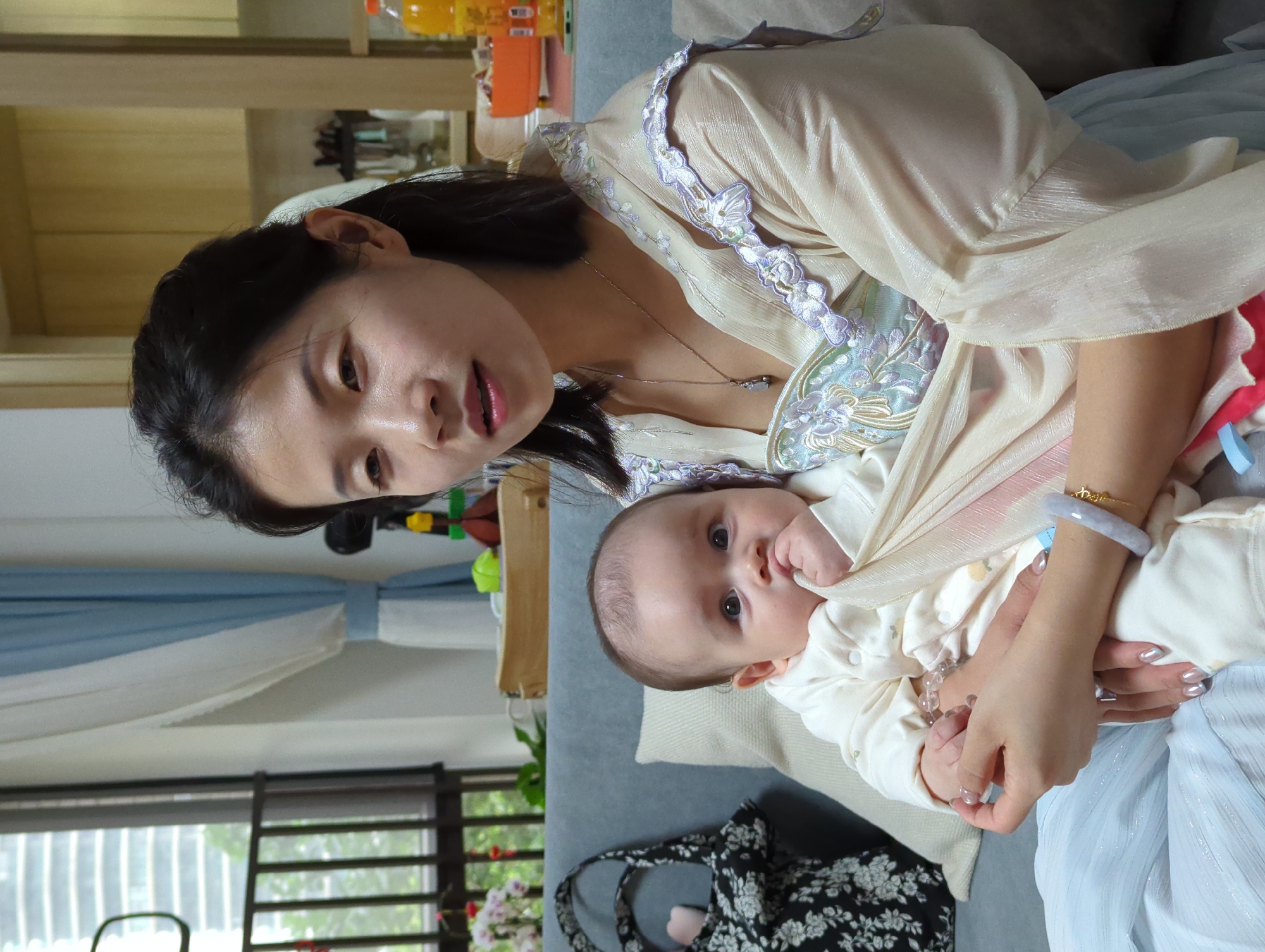 An indoor scene shows a woman holding a baby. The woman has dark hair and is wearing a light-colored top with embroidered detailing. She is looking at the camera. The baby is wearing white clothing and is partially covered by a light-colored cloth. The baby is looking at the camera. They are sitting on a grey sofa. In the background, there is a window with bars, part of a curtain, a shelf with bottles on it, a toy, and a black and white floral patterned bag. The location is potentially in the city of Shunde District, China, based on visual cues.