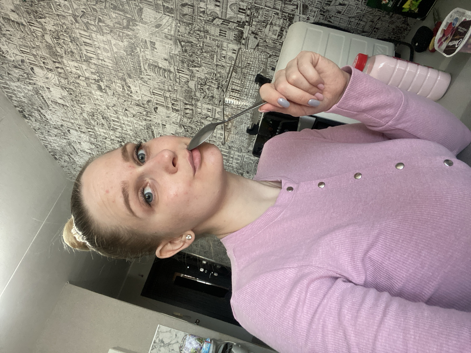 A young woman with blonde hair in a bun, wearing a pink cardigan, holds a spoon to her lips as she looks into the camera. She has blue eyes and light skin with some blemishes. The background is a wall with a patterned wallpaper depicting a cityscape. To the right, a pink bottle and a container of yogurt are visible on a surface, possibly a kitchen counter.