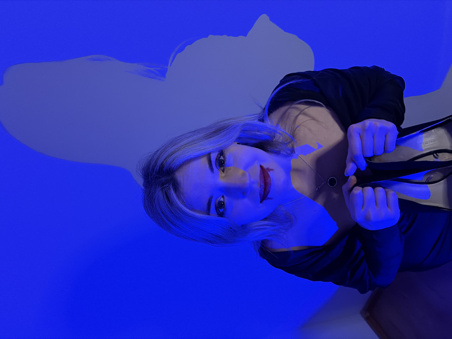 An indoor scene in Budva, Montenegro, is illuminated by a predominant blue light. A single female individual, with blonde hair and fair skin, occupies the central foreground, looking directly at the viewer. She wears dark lipstick, from which simulated blood drips from her lower lip towards her chin, suggesting a theatrical or costume theme. Her attire consists of a dark, low-cut garment and a dark jacket or shawl draped over her shoulders. A silver-colored necklace with a circular black pendant is visible around her neck. Her hands are clasped together at waist level, holding a pair of black high-heeled shoes. To the right and behind the subject, a large, indistinct shadow, consistent with a human figure, is cast upon the blue-lit wall. A light-colored wooden floor is partially visible in the bottom left corner.