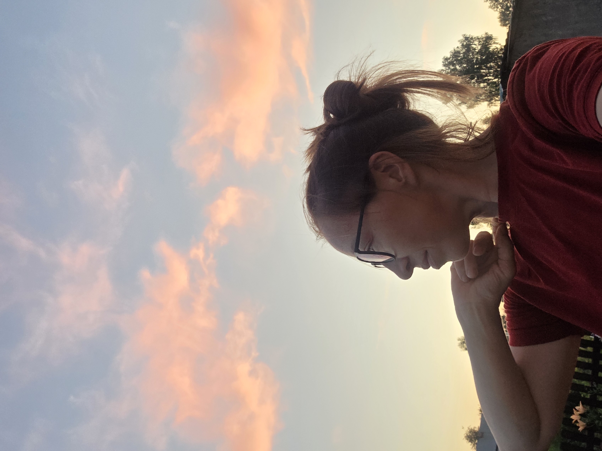 A woman with glasses and her hair in a bun, wearing a red shirt, looks to the left in profile against a sunset sky with pink clouds. The background shows trees and a fence. The scene appears to be in an outdoor setting in Буренка, Russia.