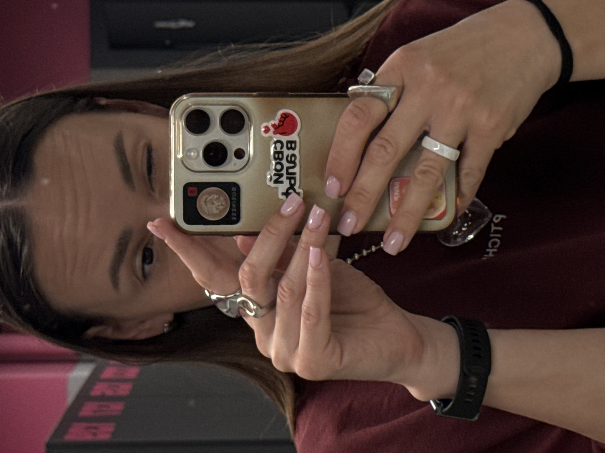 A young Caucasian woman with long, dark brown hair is taking a mirror selfie with her smartphone. She is wearing a dark red sweatshirt. On her right hand, she has a large, ornate silver ring on her index finger and a lighter silver ring on her middle finger. On her left hand, she is wearing a white ring on her middle finger. She has a black smartwatch on her left wrist and a black hair tie on her right wrist. The phone case has several stickers on it. The background appears to be a room with pink and dark gray walls, possibly in a gym or studio. There is no indication of the location being in Sochi, Russia.