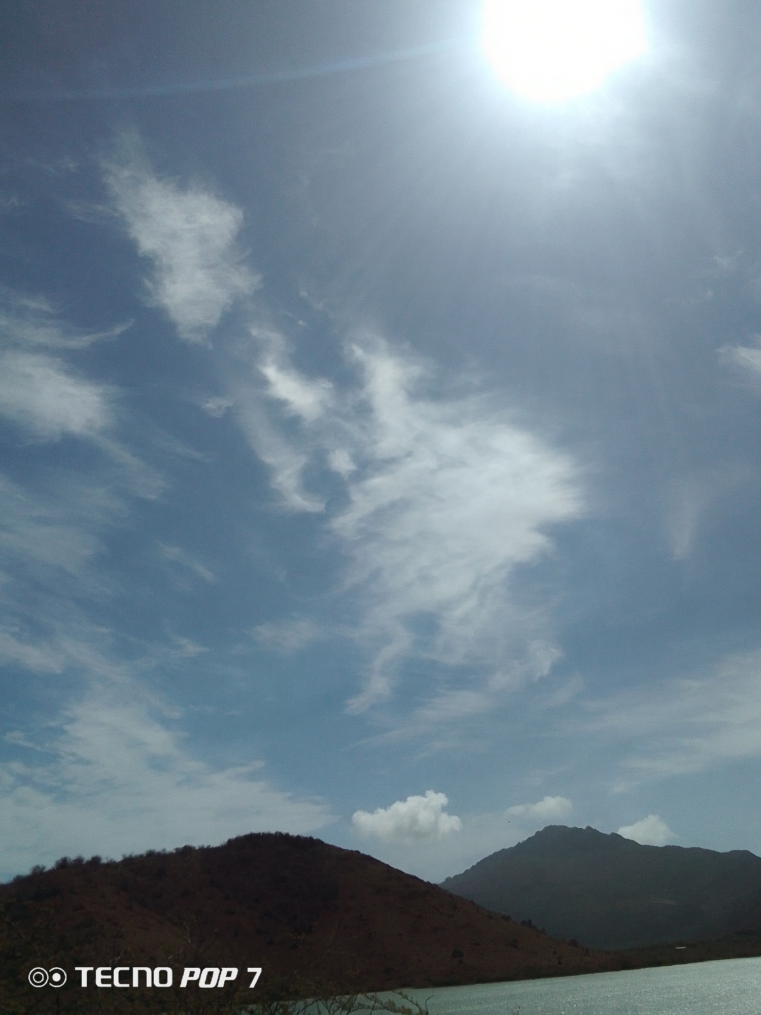 The image shows a landscape with two visible hills and a body of water, likely a bay or large lake, in the foreground. Above is a bright, overcast sky with thin, wispy clouds and a prominent sun. The text "TECNO POP 7" is at the bottom of the image, indicating the device used to take the picture. Given the prompt details, this image is presumably taken in Pedro González, Venezuela.