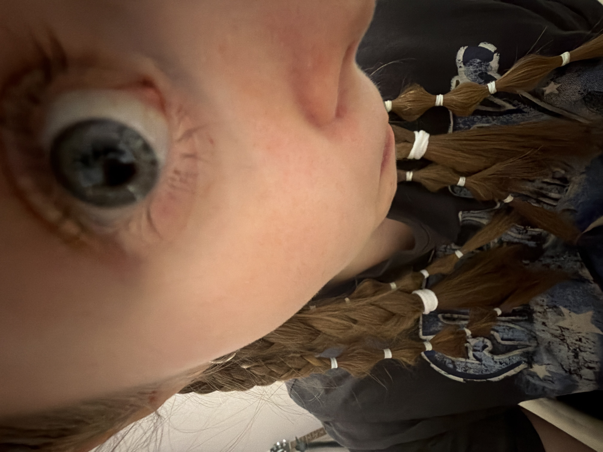 This close-up portrait captures a person from an unconventional, tilted angle, focusing predominantly on their face and hair. The subject has fair skin, with a prominent blue-grey eye wide open and appearing focused. Visible eyelashes frame the eye, and parts of the nose and cheek are also discernable. Their long, brown hair is styled into numerous small braids, each segment secured by white elastic bands, creating a segmented appearance. These braids rest against a dark-colored t-shirt or garment that features a faded blue and white graphic design, possibly incorporating stars or abstract patterns.
The setting appears to be indoors, suggesting a casual environment like a home. In the lower background of the original image, a faint outline possibly indicates part of a musical instrument, such as a guitar neck. The time of day and weather conditions cannot be determined from the image. Due to the extreme close-up and partial view of the face, no specific emotions are clearly conveyed. There is no visible text in the image. While the location context provided is Perm, Russia, this is not visibly depicted within the photographic content itself. The image seems to be a personal, possibly self-taken, photograph.