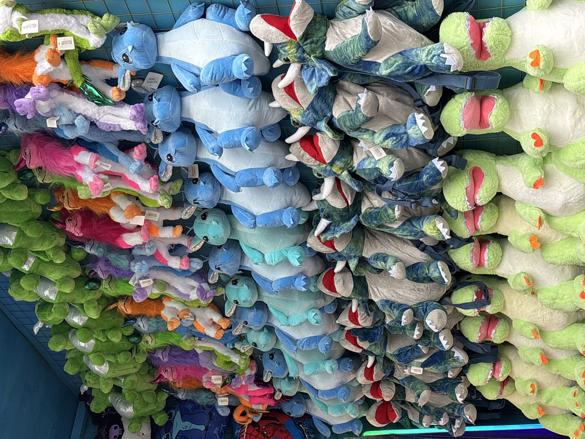 The image shows a display of stuffed animals hanging from a grid, likely in a retail setting. The animals are arranged in rows and columns, creating a dense, colorful assortment. There are several types of animals: green turtles, blue creatures, triceratops with red head plates, and pink, purple, and orange unicorns. The animals are suspended from the grid by small straps attached to their bodies. The location is not Izhevsk, Russia.