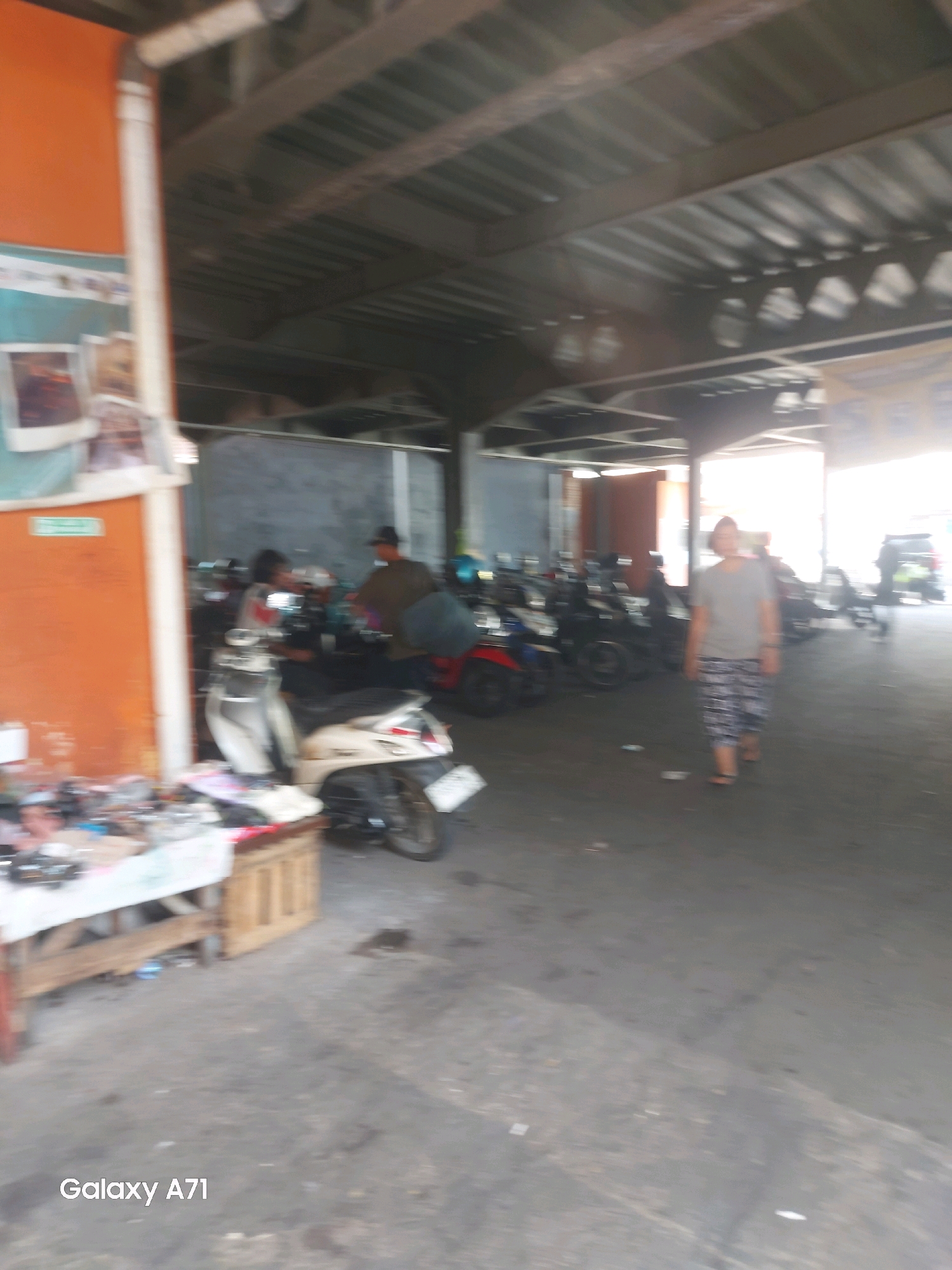 The image depicts an outdoor market stall area in Makassar, Indonesia, with a focus on a row of parked motorcycles. A person, possibly a vendor, is seen behind a table laden with goods. In the background, other individuals are present, and several more motorcycles are parked. The environment suggests a covered area, likely a marketplace, with a corrugated metal roof overhead supported by beams. The ground is paved, and some debris is visible. A person in the foreground walks away from the camera, wearing patterned shorts and a grey t-shirt. The image is branded with "Galaxy A71" in the lower left corner.