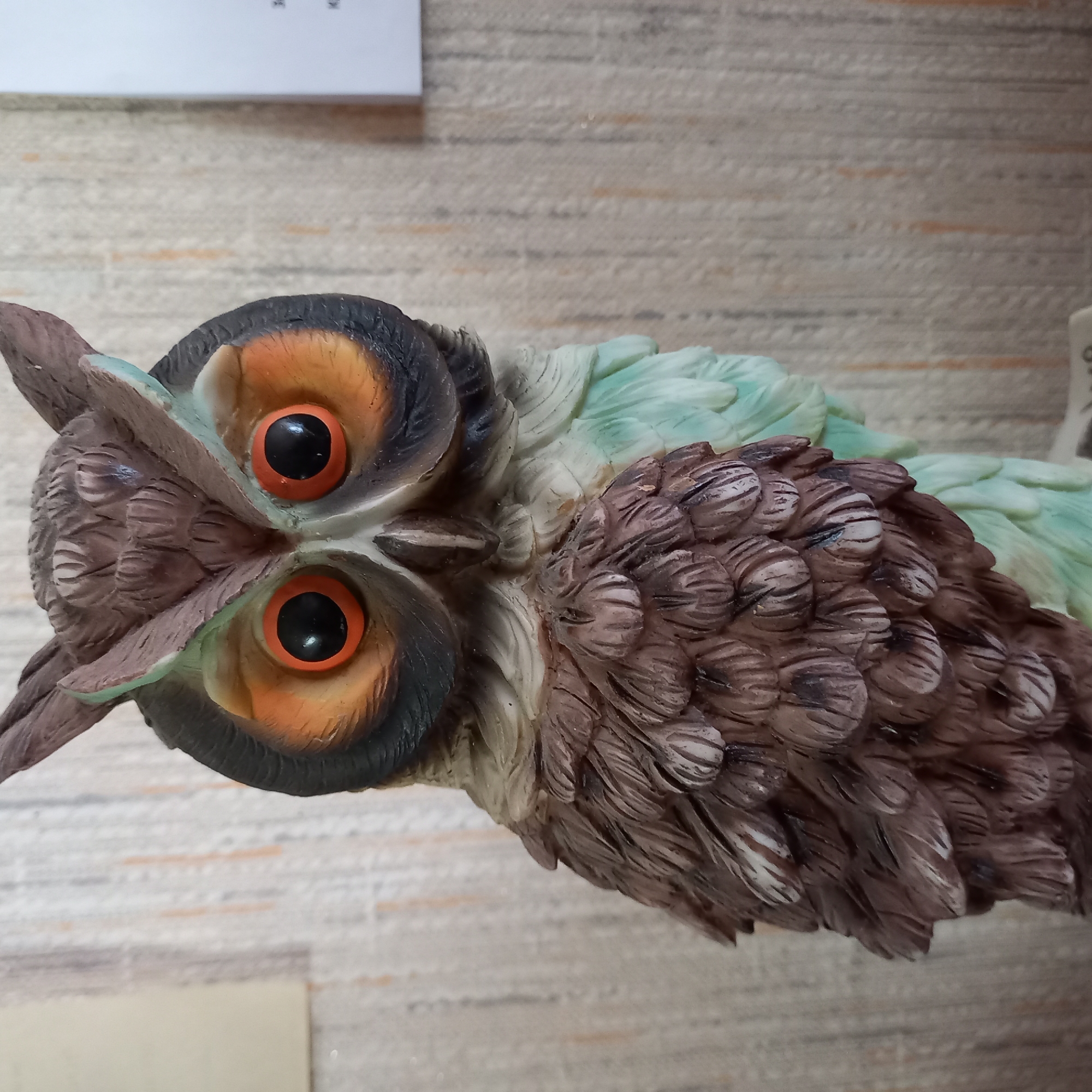 A close-up, eye-level, portrait-oriented photograph shows a highly detailed figurine of an owl. The owl figurine is positioned in the center of the frame, facing forward. It is depicted with large, round, black eyes surrounded by bright orange irises. The owl's head features prominent ear tufts and is colored in shades of brown and black, with green accents around the eyes and wingtips. The body of the owl is textured to resemble feathers, with intricate detailing in brown and light green. The background is a textured wall, appearing to be wallpaper, with vertical stripes of beige and subtle orange and grey lines. To the right of the owl, a white paper is visible attached to the wall, with some faint black text or markings on it. The lighting is even, casting no harsh shadows, suggesting it may be indoors with artificial light. The overall impression is that of a decorative object. The image is in focus, showcasing the fine craftsmanship of the figurine. The specific location in the city of Unknown, Ukraine cannot be determined from the visual information provided in the image.