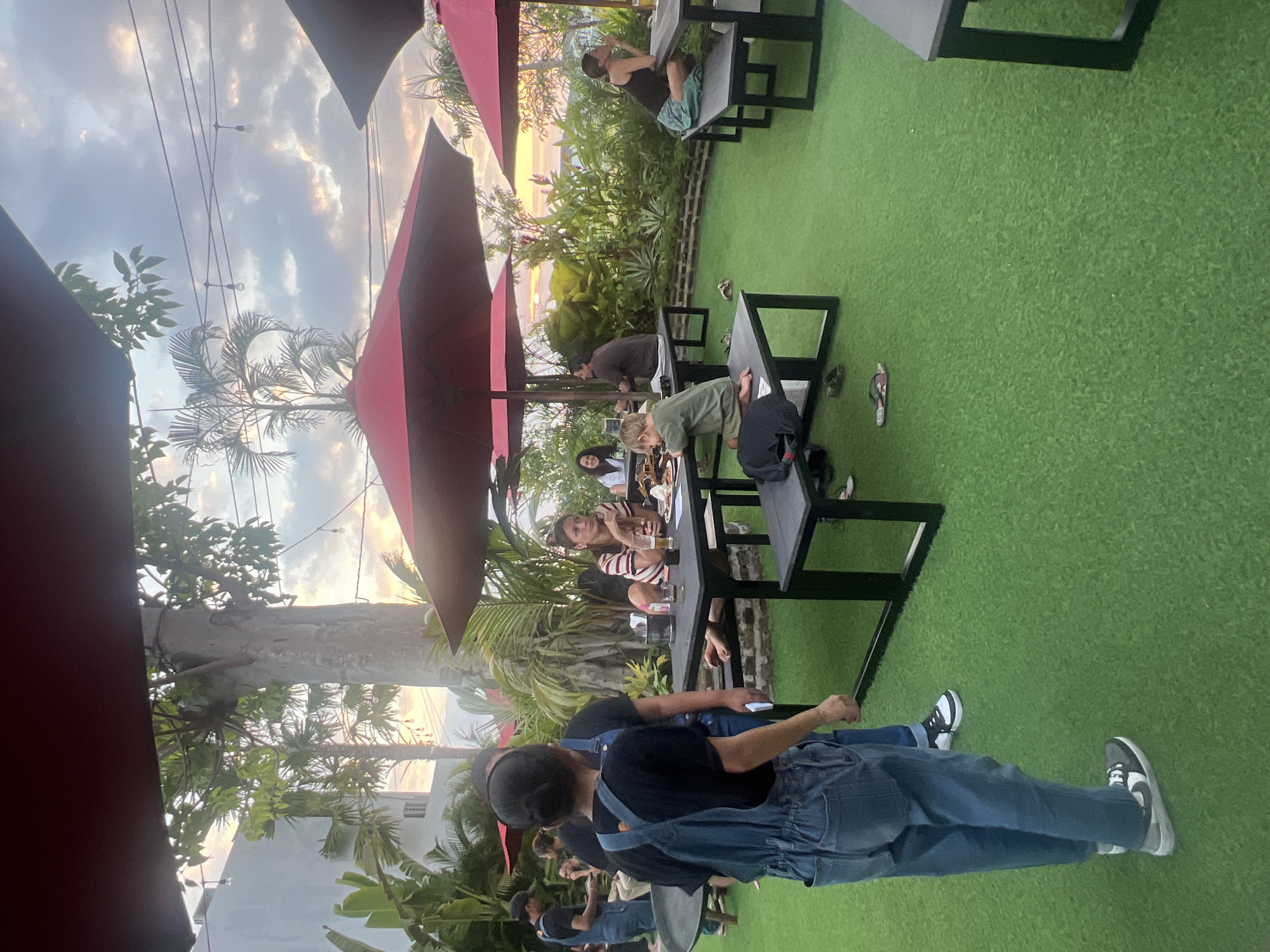 This image depicts an outdoor dining area with an artificial turf floor, illuminated by string lights and under the shade of red umbrellas. Several picnic tables with benches are scattered across the area, occupied by patrons. In the foreground, two individuals are visible, one wearing denim overalls and a black t-shirt, and the other in a black t-shirt and denim jeans. The background reveals a lush green landscape with tropical plants and trees, suggesting a warm climate. The sky is filled with clouds, hinting at either sunrise or sunset. The overall ambiance is casual and relaxed, consistent with an outdoor restaurant or cafe setting. There is no visible indication or context that suggests this scene is located in the city of Kediri, Indonesia.