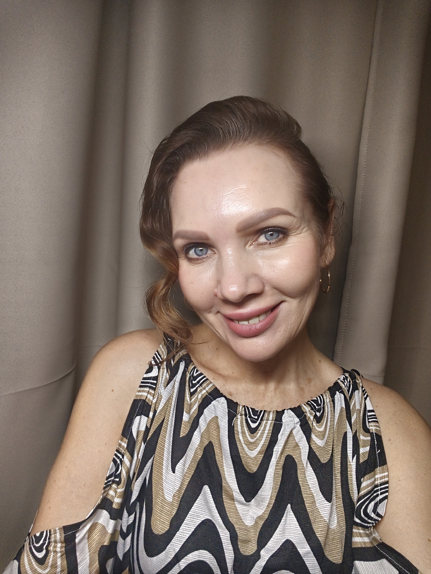A woman with fair skin, blue eyes, and reddish-brown hair is smiling at the camera. She is wearing a sleeveless top with a wavy pattern of black, white, and tan. The background is a plain, neutral-colored curtain. The location is Unknown, Unknown.