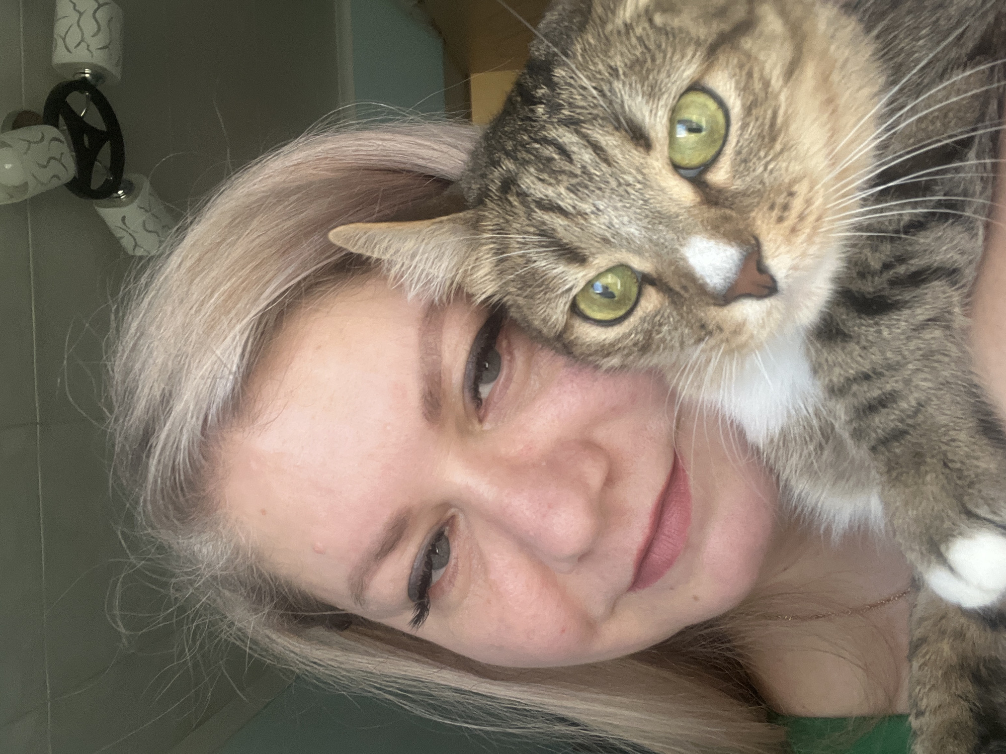 A woman is holding a tabby cat. The woman has blonde hair and is wearing makeup. The cat has green eyes and a white nose. The background is blurry, but appears to be a room with a light fixture. There is no information to suggest that the location is Izhevsk, Russia.