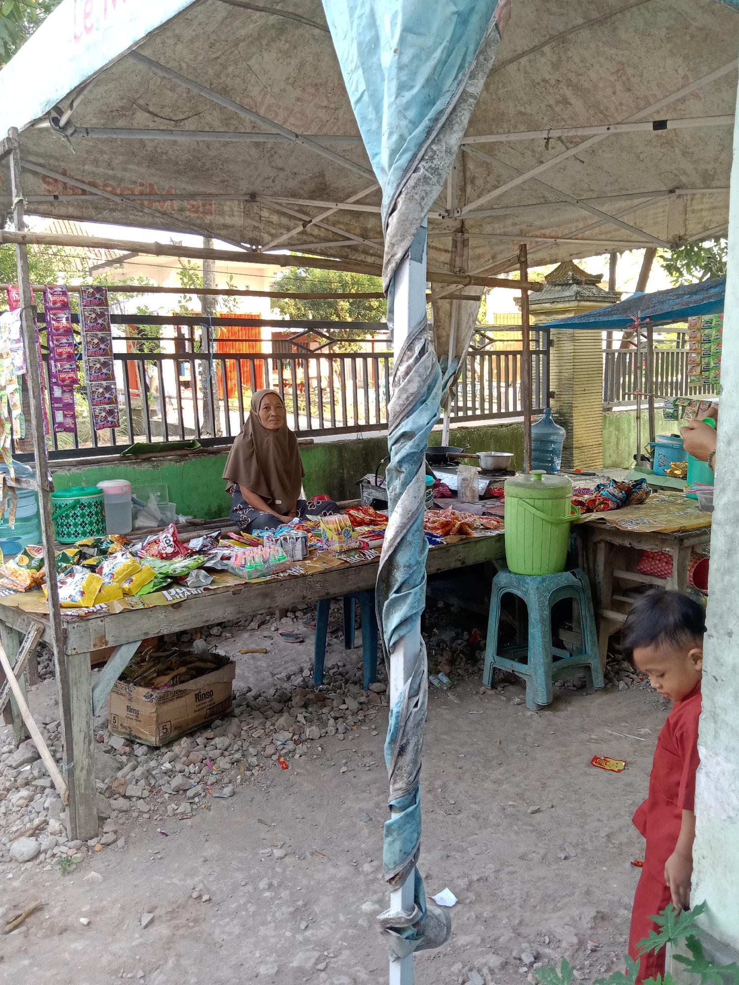 In Sugihwaras, Indonesia, an outdoor stall operates under a makeshift canopy supported by metal poles. A woman wearing a brown hijab and patterned clothing sits behind a wooden table laden with various snacks and drinks, including packaged chips, candy, and bottled beverages. To the right, a young boy in a red shirt stands partially obscured by a white wall, looking down. The ground is covered in dirt and scattered stones. In the background, a wooden fence and some buildings are visible.