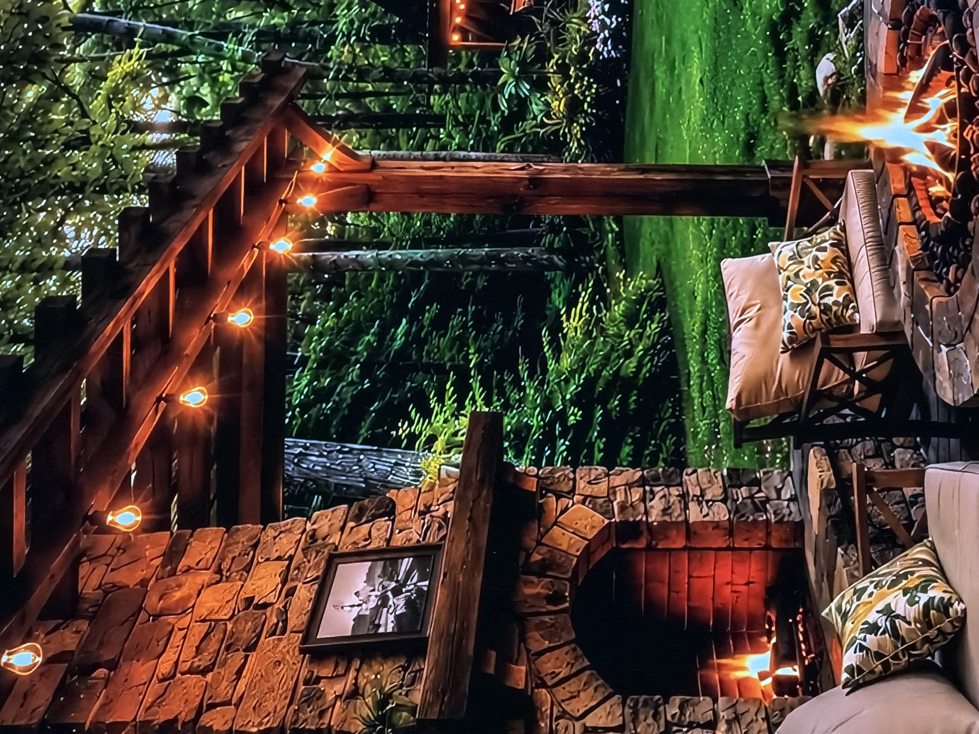 This media file captures a cozy outdoor living space, likely a backyard or garden area in Sofiivska Borshchahivka, Ukraine, during the evening or night. The scene is dominated by warm, artificial lighting from string lights and a roaring fire, creating an inviting atmosphere.
In the foreground, a robust, rustic stone wall serves as the base for a fireplace or fire pit, where bright orange and yellow flames are actively burning. A small, framed picture with an unidentifiable monochrome image hangs on this stone wall. To the right of the fire, comfortable outdoor seating is visible, featuring thick, light-colored cushions and decorative pillows, one adorned with a green and yellow leaf pattern, suggesting a place for relaxation.
Above and to the left, a wooden pergola or deck structure is prominent, with dark wooden beams and slats. A string of exposed bulb lights hangs from this structure, casting a warm glow across the scene and adding to the ambiance. A large, horizontal wooden beam extends across the middle of the frame, further contributing to the rustic aesthetic.
The background is filled with lush, vibrant green foliage, trees, and grass, indicating a verdant natural setting surrounding the constructed space. The overall impression is one of comfort, warmth, and peaceful outdoor enjoyment. No discernible text is visible in the image.
