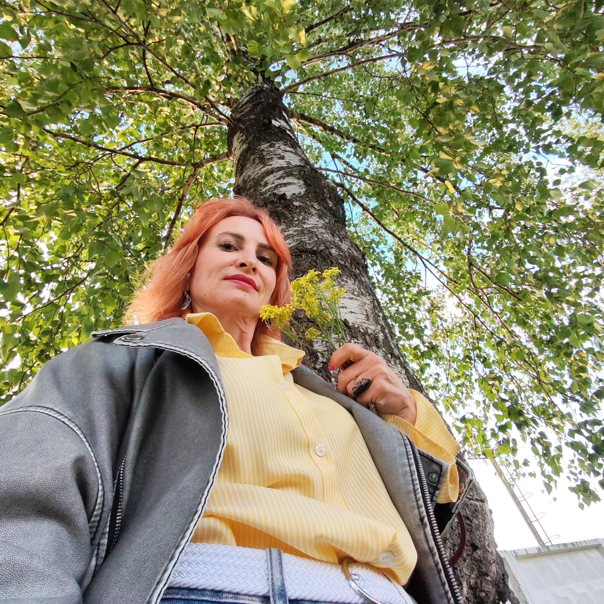 A fair-skinned woman with reddish hair stands beneath a birch tree in an outdoor setting. She wears a grey jacket over a yellow-and-white striped shirt, and blue jeans with a white belt. She holds a small bunch of yellow flowers in her right hand. The camera angle is from below, looking up at her and the tree. Green leaves fill the upper half of the frame. The location is Chaikovsky, Russia. In the background, a ladder leaning against the side of a white structure is visible.