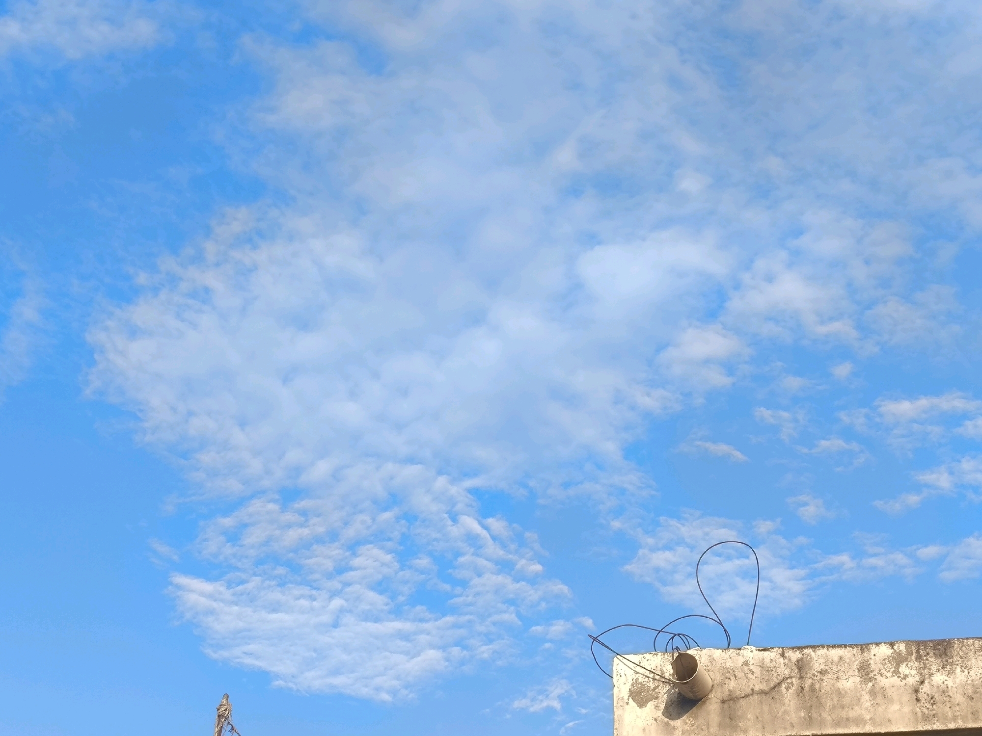 A low-angle, medium shot captures a concrete rooftop in Chhajpur Kalan, India, against a vibrant blue sky filled with scattered, wispy white clouds. The rooftop's surface is textured and weathered, showing patches of discoloration and wear. Protruding from the rooftop's edge is a circular, grey pipe, likely a drain or vent. Several black electrical wires are visible, one forming a loop against the sky and another extending downwards. In the background, to the left, a thin, vertical structure, possibly a pole or antenna, is partially visible against the sky.
