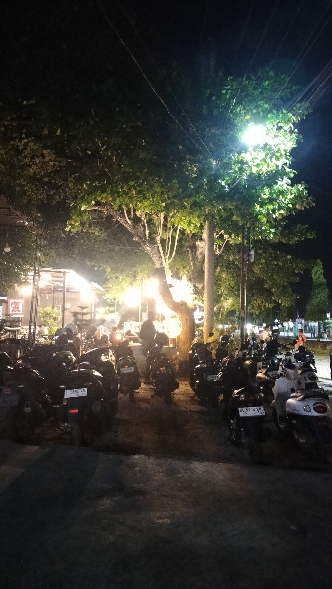 The image depicts a nighttime scene in Lhokseumawe, Indonesia, likely in a commercial or recreational area. A large tree with dense foliage dominates the upper portion of the frame, illuminated by a bright street lamp attached to its trunk. The ground below is covered with parked motorcycles, suggesting a public gathering or establishment. In the background, a building with open-air seating and warm lighting indicates a cafe or restaurant. Silhouettes of people are visible in the dimly lit interior of the establishment and standing near the motorcycles. The overall atmosphere is dark, with the artificial lights creating pools of illumination amidst the shadows. License plates are visible on some of the motorcycles, including "BL 6685 NAU" and "BL 5174 KRJ".