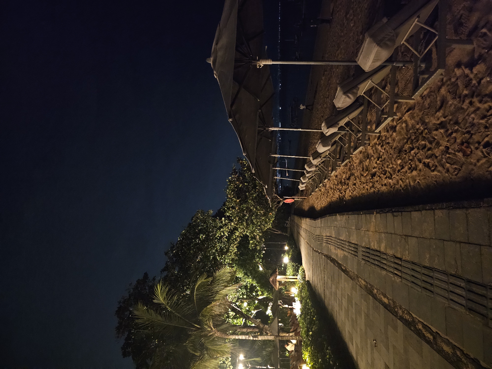At night in Denpasar, Indonesia, a paved path runs alongside a sandy beach. Several beach umbrellas and chairs line the sand. Trees and shrubs border the path, illuminated by artificial lights. The sky is dark.