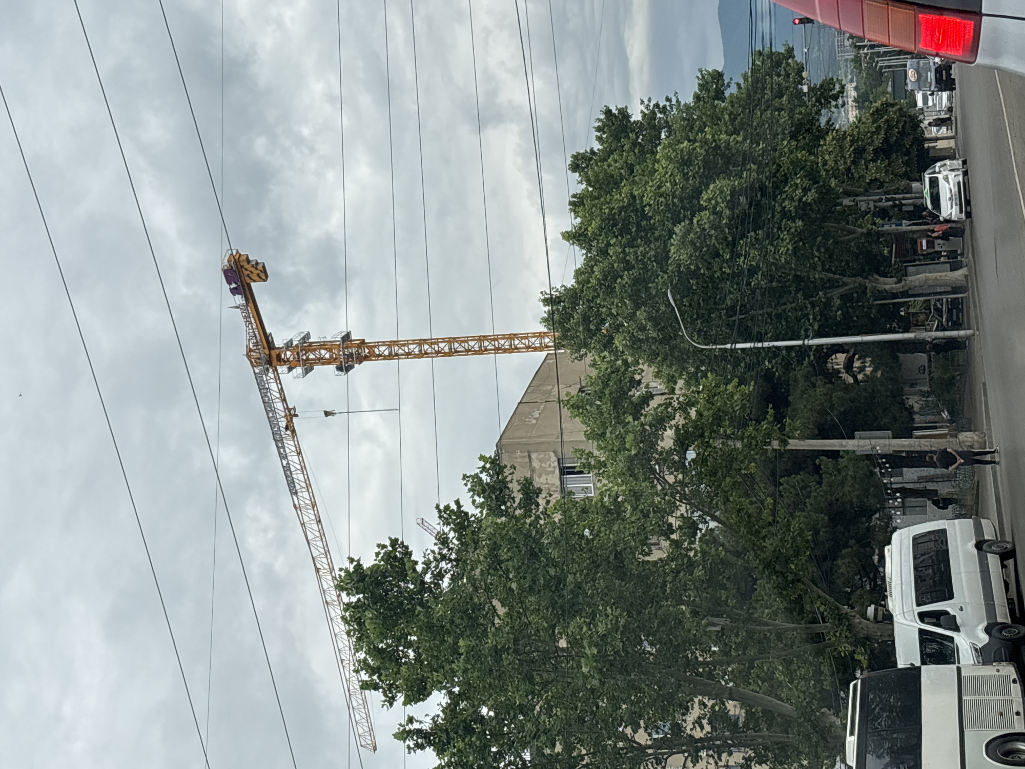 The image captures a street scene in Tbilisi, Georgia, dominated by a large yellow construction crane extending across the sky. The crane is positioned behind a cluster of lush green trees, with a building that appears to be under construction or renovation visible behind the foliage. The sky is overcast with heavy clouds, suggesting a cloudy or possibly rainy day.
In the foreground, several vehicles are present. A white bus is parked on the left side of the frame, and a white minivan is positioned next to it. Further down the street, a white car is driving, and other vehicles are partially visible, including a dark-colored truck and the rear of a car with its brake lights illuminated.
The street itself is paved, with a sidewalk running alongside it. Several streetlights and power lines crisscross the scene, adding to the urban environment. A few individuals are visible on the sidewalk, including a person standing with their hands behind their back and another individual walking. The presence of the construction crane, the building, and the vehicles indicates an active urban area undergoing development. The overall atmosphere is muted due to the cloudy weather.