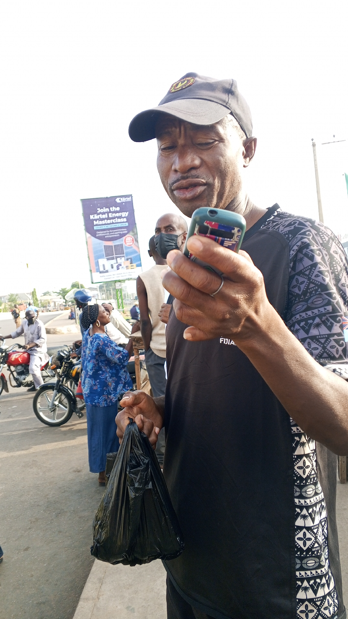 A man in a black patterned shirt and baseball cap is holding a black plastic bag and a cell phone near a busy road in Maiburiji, Nigeria. Other people stand nearby, including one wearing a face mask. Motorcycles are parked along the roadside. A billboard advertising the "Kartel Energy Masterclass" is visible in the background.