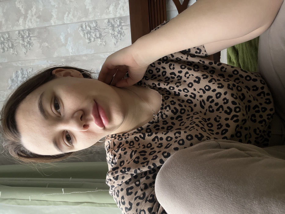 This image depicts a single female individual within an indoor residential setting in Leninakan, Russia. The subject, a woman with dark hair pulled back from her face, is positioned in a close-up, slightly low-angle view, facing directly towards the camera. She has light skin, dark eyebrows, and visible light lip gloss. Her left hand is raised, with fingers gently resting on her lower jaw and neck on the right side of the frame. She wears a tan-colored short-sleeved top with a black leopard print pattern and light beige or grey trousers, with her right knee bent and visible in the lower left foreground. A delicate silver-colored necklace with a small pendant is visible around her neck, and a small hoop earring is present in her left ear.
The background shows a light green sheer curtain on the left, indicative of a window. The wall behind the subject is covered with wallpaper featuring a light grey or white background and a recurring pattern of stylized dark trees or cloud-like formations. To the right, part of a dark wooden furniture piece, possibly a headboard or chair frame with horizontal slats, is visible. A dark green fabric or blanket is partially visible in the lower right corner of the frame. The lighting appears to be natural, originating from the left.