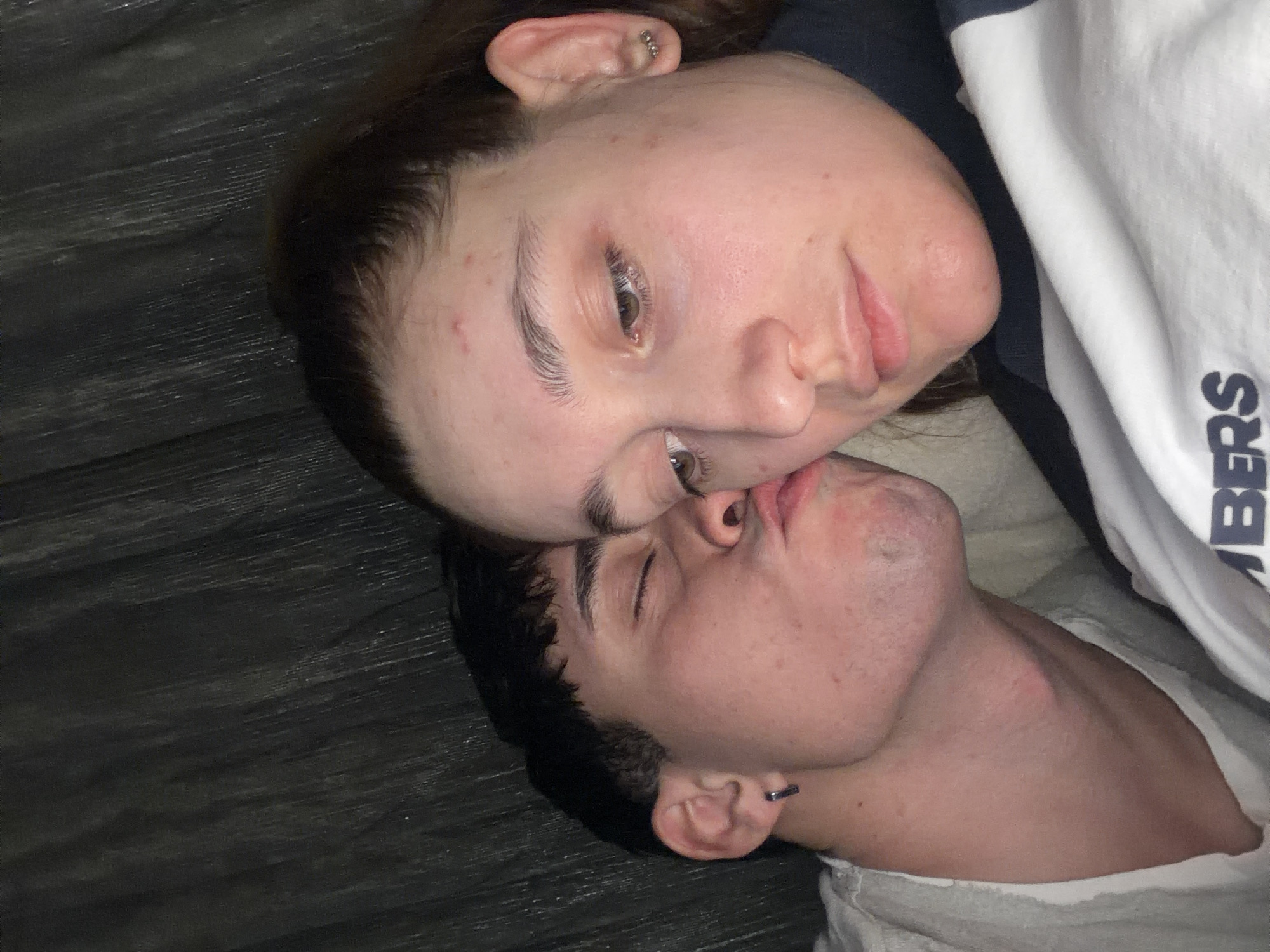 A photograph shows two individuals, a woman and a man, lying down with their faces close together. The woman is positioned at the top of the frame, with her head tilted slightly. She has light skin, brown hair, and an earring in her ear. The man is positioned beneath her, with his eyes closed. He has dark hair and a piercing in his ear. They appear to be close and intimate. The background consists of a dark wooden surface and a white garment with the letters "BERS" visible. The location is not identifiable as Blagoevgrad, Bulgaria.