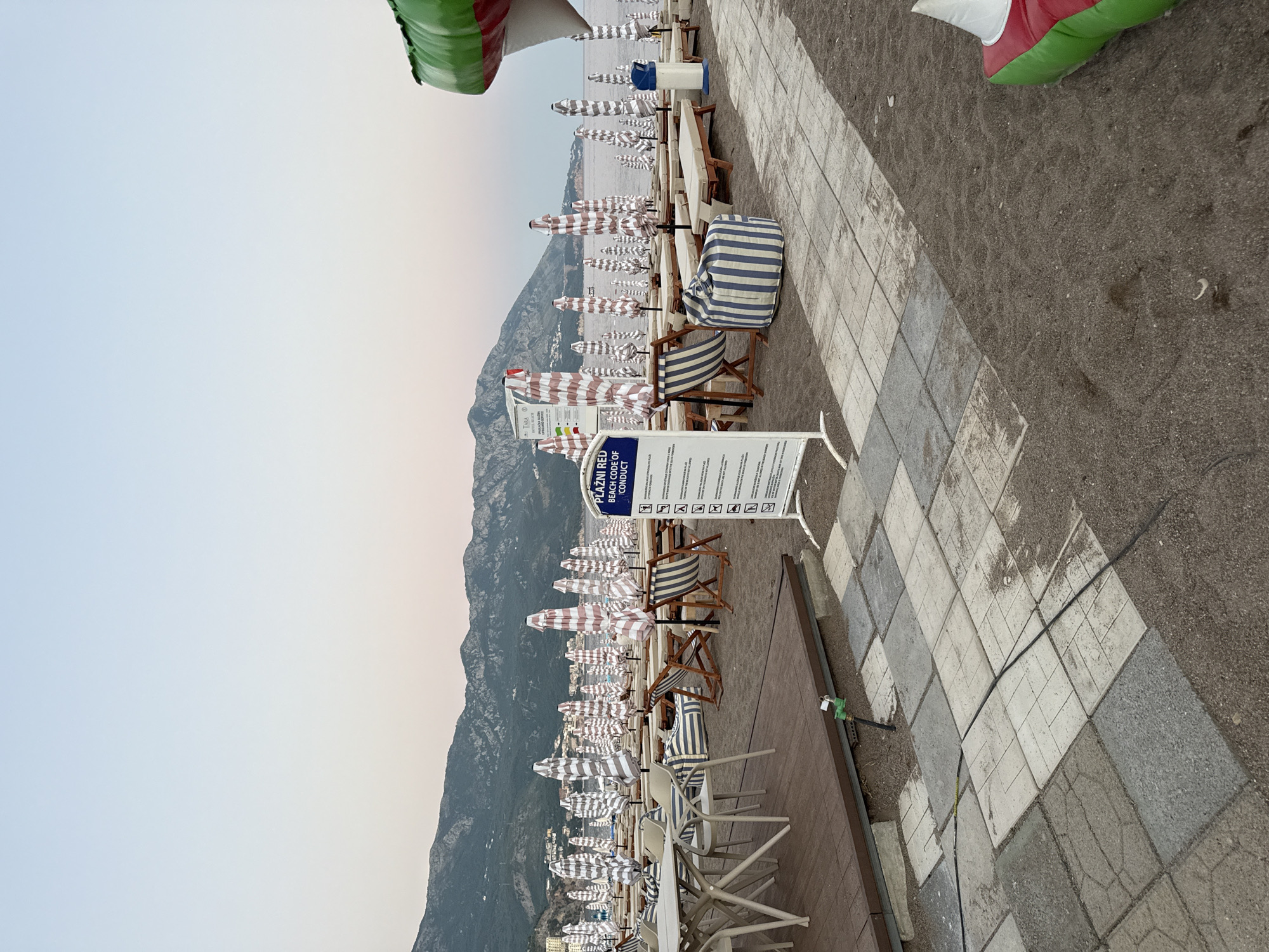 The image depicts a beach scene in Bečići, Montenegro. The beach is lined with numerous closed umbrellas and empty lounge chairs arranged in rows, suggesting it is early morning or late evening. A sign with rules and regulations, written in a foreign language, is prominently displayed in the foreground. To the left, a wooden deck area with tables and chairs is visible. In the background, mountains form a scenic backdrop to the coastline. The sky is clear with a gradient of light blue to pale pink. The sand appears dark, and the sea is calm. There are no people visible in the frame.