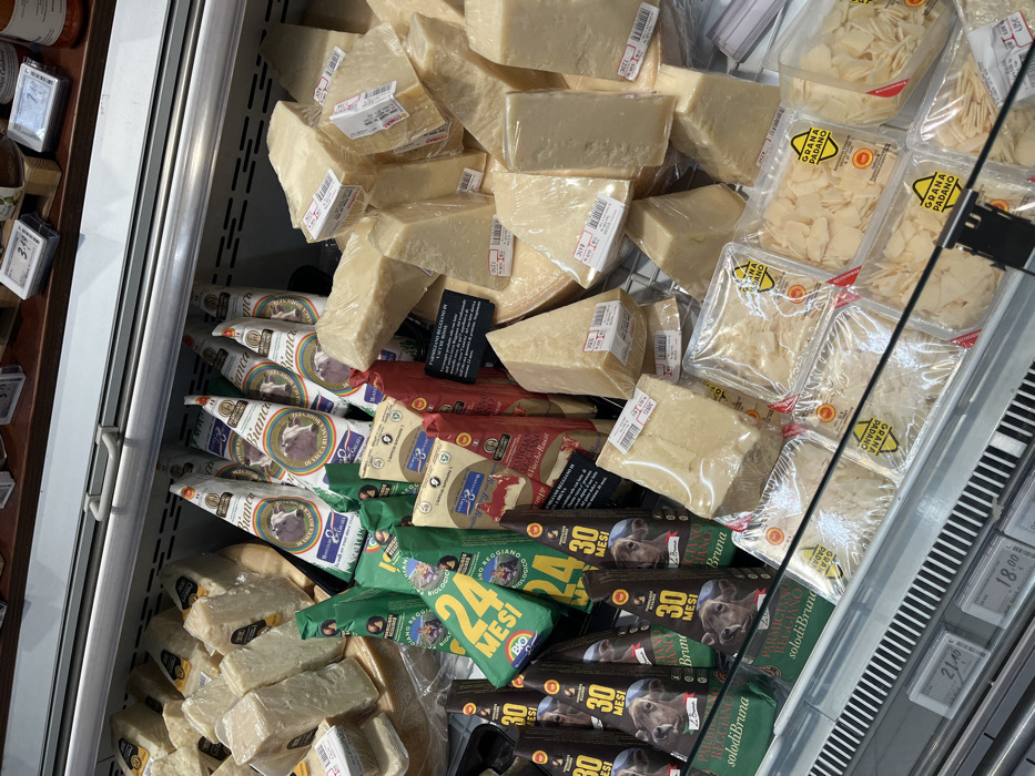 This image displays a refrigerated deli counter section in a grocery store in Monsummano Terme, Italy, dedicated primarily to various Italian cheeses. The brightly lit display case is filled with a dense assortment of cheese products, arranged on tiered shelves and behind a glass barrier.
The majority of the visible cheese consists of irregular, pale yellow blocks and wedges, typical of hard aged cheeses, individually wrapped in clear plastic film and bearing white adhesive price labels.
Behind these unwrapped portions, several types of pre-packaged cheeses are visible. These include:
* Triangularly packaged portions of "PARMIGIANO REGGIANO BIOLOGICO" (Organic Parmigiano Reggiano) in green packaging, some marked "24 MESI" (24 months).
* Triangularly packaged portions of "PARMIGIANO REGGIANO Bruna" in dark brown/black packaging, featuring an image of a calf and indicating "30 MESI" (30 months).
* White, triangularly packaged portions labeled "MONTECCHIO MONTONE DELLA MUCCA BIANCA" (Montecchio Lamb from White Cow) with a cow head logo.
* Red/burgundy packaged portions of "PARMIGIANO REGGIANO Delle Vacche Rosse" (Parmigiano Reggiano from Red Cows).
At the bottom of the display, multiple clear rectangular plastic containers are filled with pre-shredded or shaved cheese, each labeled "GRANA PADANO." A metal grate forms the base of the display, and digital price tags are visible on a rail beneath the products. The upper section of the display, partially visible, shows additional packaged items and price labels on a dark shelf. No individuals are present in the scene.