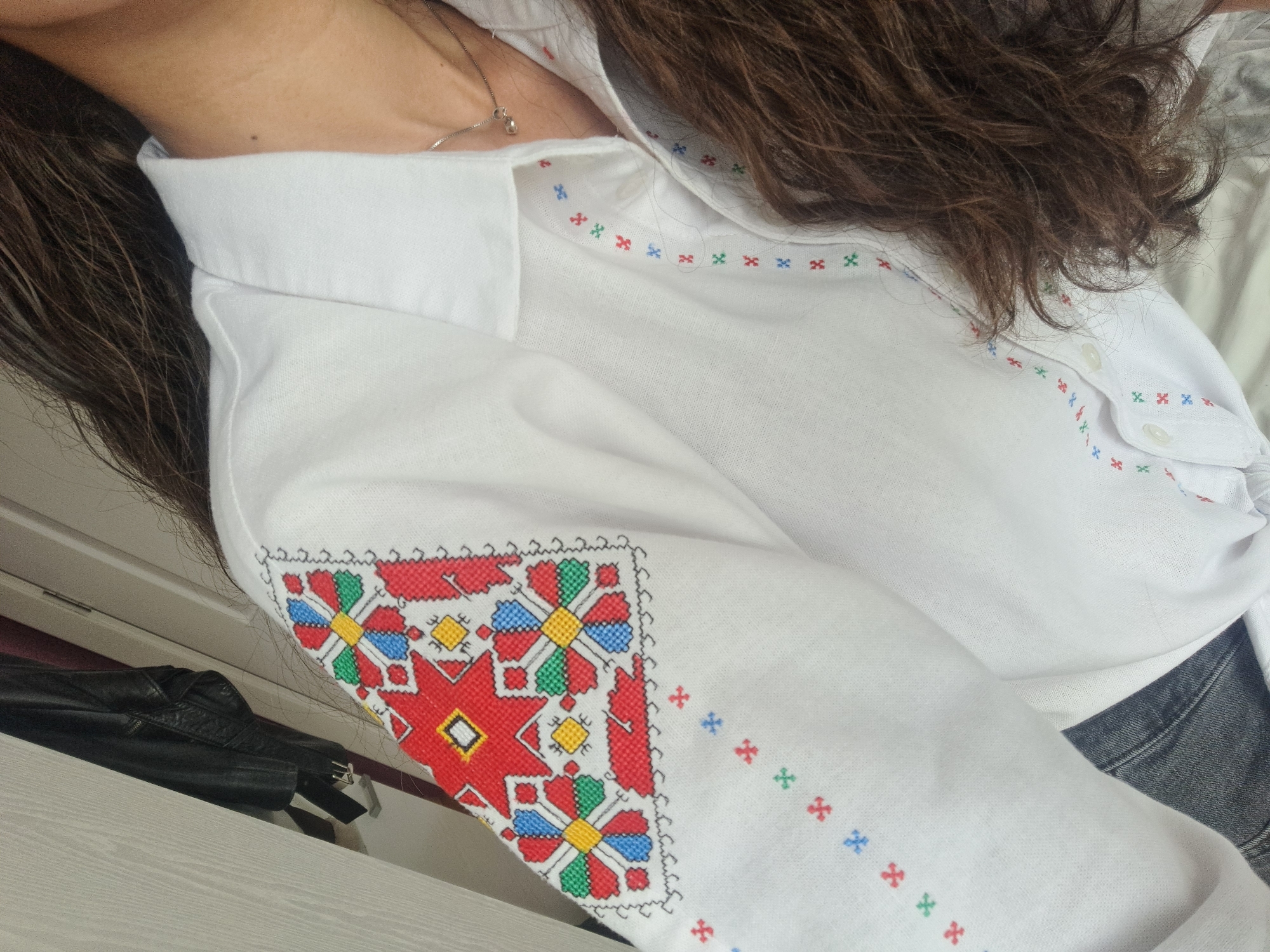 A close-up shot focuses on a person wearing a white shirt with embroidered patterns on the sleeve. The embroidery features red, blue, green, and yellow geometric shapes. The person is also wearing a silver necklace. A portion of their brown hair is visible. A black leather jacket is partially seen on a light-colored surface.