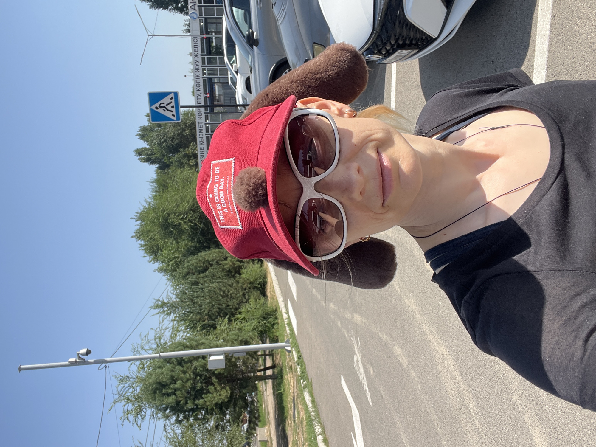 A woman, wearing a red cap with floppy brown ear-like attachments and white-rimmed sunglasses, stands facing the camera in a parking lot. The cap has a patch with the text "THIS IS GOING TO BE A GOOD DAY." She is wearing a black t-shirt and a thin dark necklace. Behind her, cars are parked in a lot adjacent to a paved road with white lane markings. In the background, a building with signage in Cyrillic script, a pedestrian crossing sign, trees, and a lamppost with a security camera are visible. The sky is clear and blue, indicating it is daytime. The signage on the building appears to be in Kazakh and possibly Russian, which is consistent with the city of Almaty, Kazakhstan.