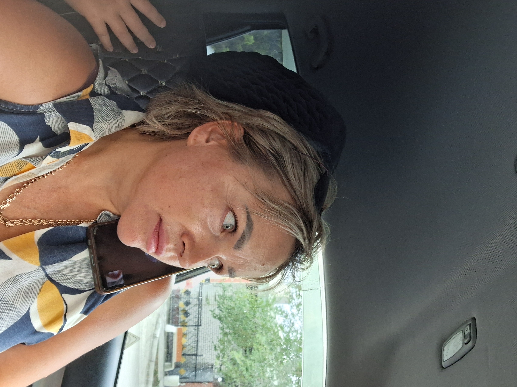 A woman with blonde hair is in the passenger seat of a car, holding a smartphone to her ear. She is wearing a patterned top and a gold chain necklace. The interior of the car is visible, including the headrest and ceiling. Through the car window, a residential area with trees and a fence can be seen. The environment suggests a daytime scene in the city of Semey, Kazakhstan.