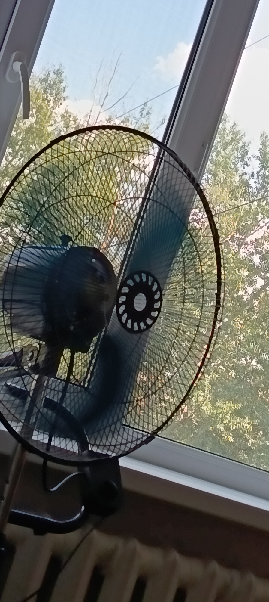 A close-up, low-angle, vertical shot shows a black oscillating fan positioned in front of a window. The fan's grille is dark and circular, with a diamond pattern. Part of the fan's motor housing is visible, and one of its blue blades is blurred, indicating movement. The window behind the fan reveals a bright, slightly cloudy sky and the green foliage of trees. To the left of the fan, a white window latch is visible. The bottom of the image shows a section of a light brown radiator. The scene appears to be indoors, with the fan facing the window. There are no people in the image. The location in Qarağandı city administration, Kazakhstan is not determinable from the visual information provided.