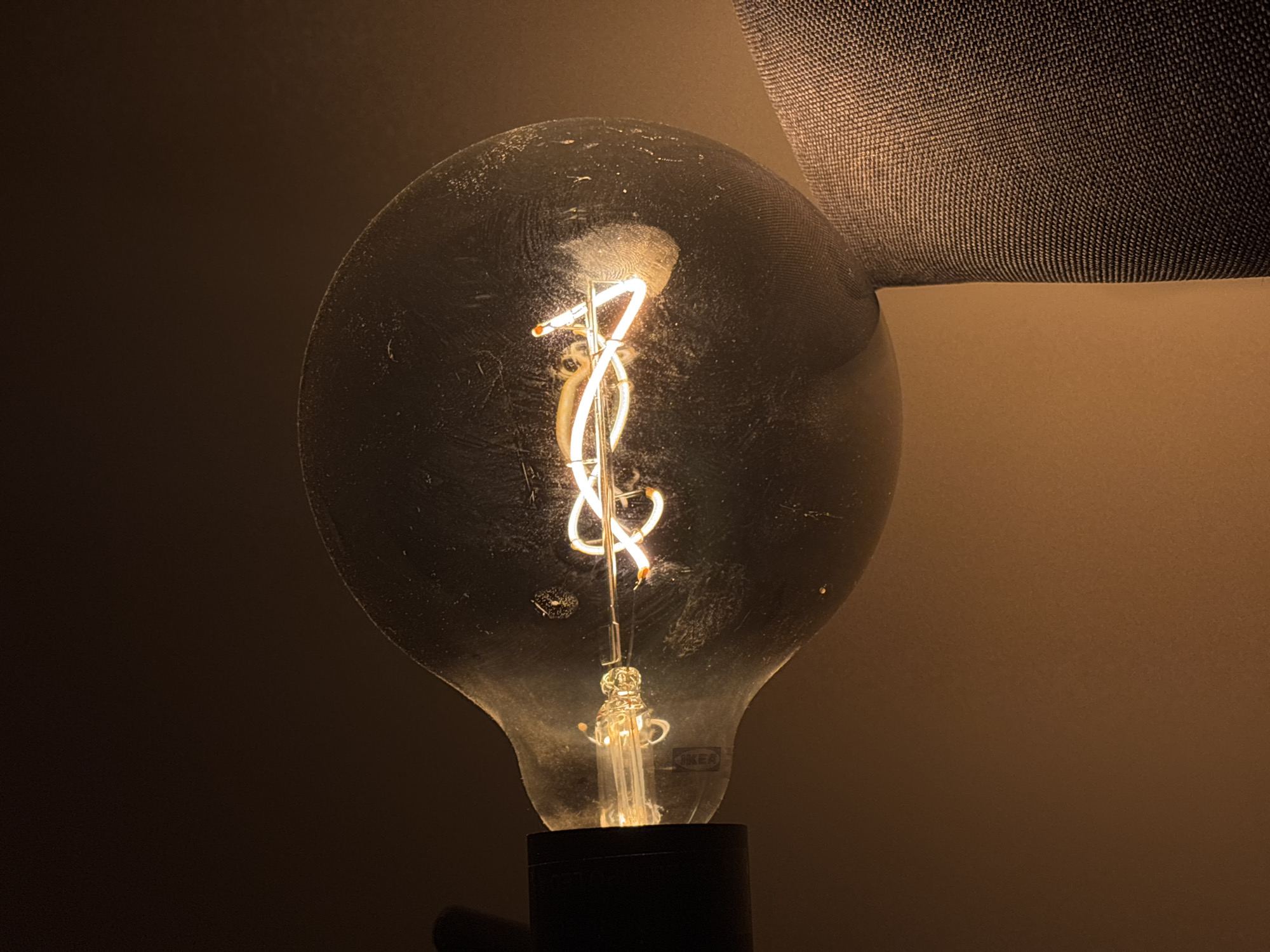 A dimly lit, close-up shot features a vintage-style light bulb with a visible filament, possibly located in Santa Clara Ocoyucan, Mexico. The bulb's glass is slightly dusty, and it's mounted on a black base. A portion of a lamp shade is visible on the right side of the bulb. The background is a muted brown color, suggesting an interior setting. No people or interactions are visible in the image.